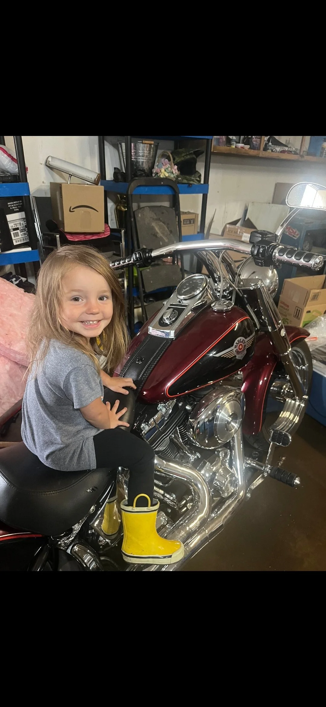Young girl with yellow rain boots, gray shirt, and black pants sitting on a shiny black and red motorcycle in a garage surrounded by shelves, boxes, and tools.