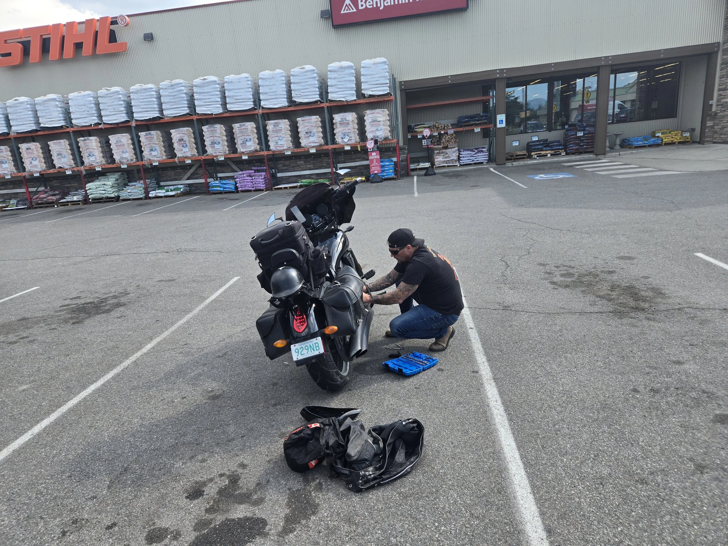 A man with tattoos, wearing a black cap, black t-shirt, and jeans, kneeling next to a black motorcycle in a parking lot outside a store. The man appears to be working on the motorcycle. There are bags and a blue toolbox on the ground near him. The st