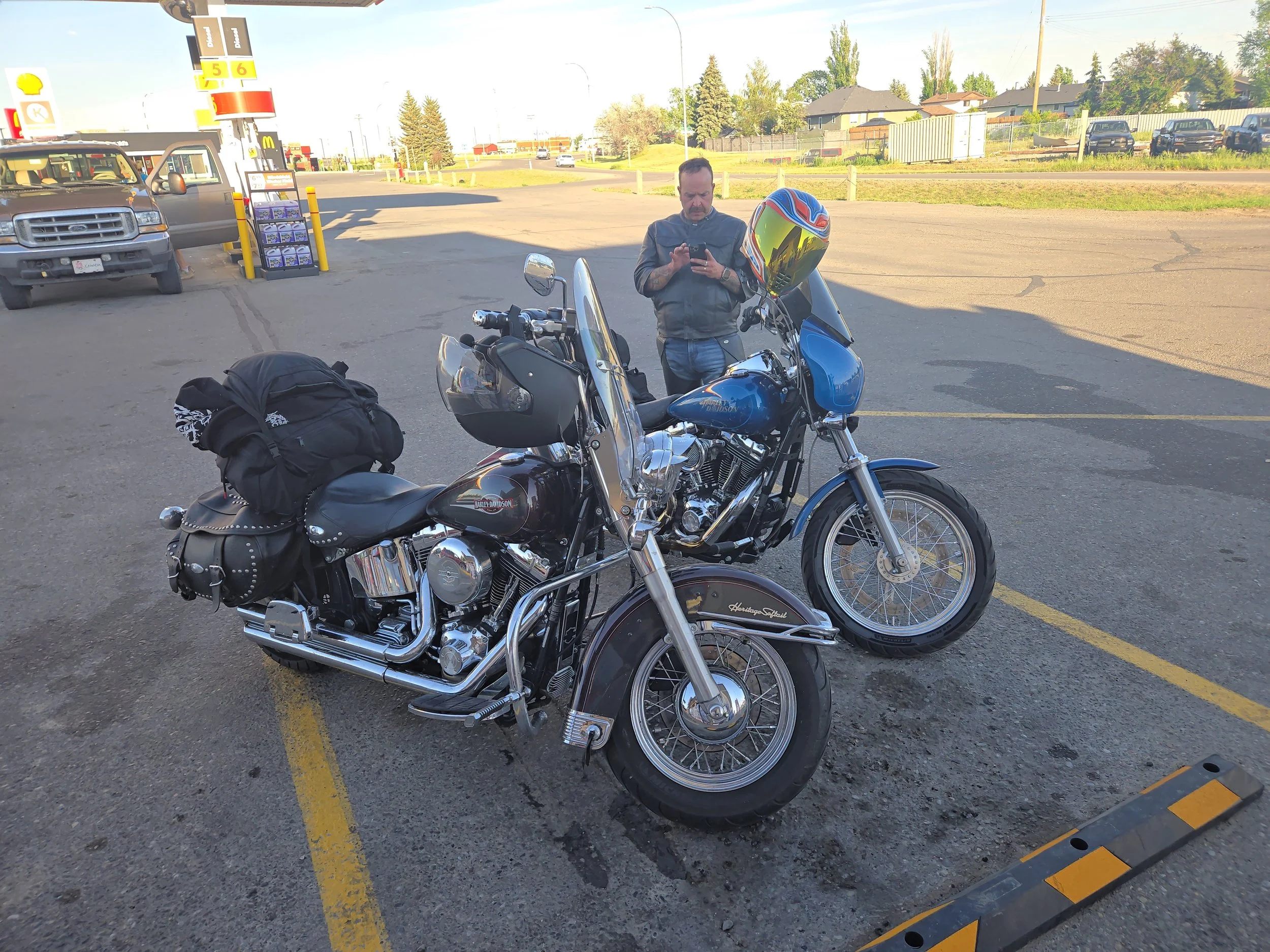 Two motorcycles parked in a parking lot near a gas station, one with a rider and a man standing beside it checking his phone, with houses and vehicles in the background.