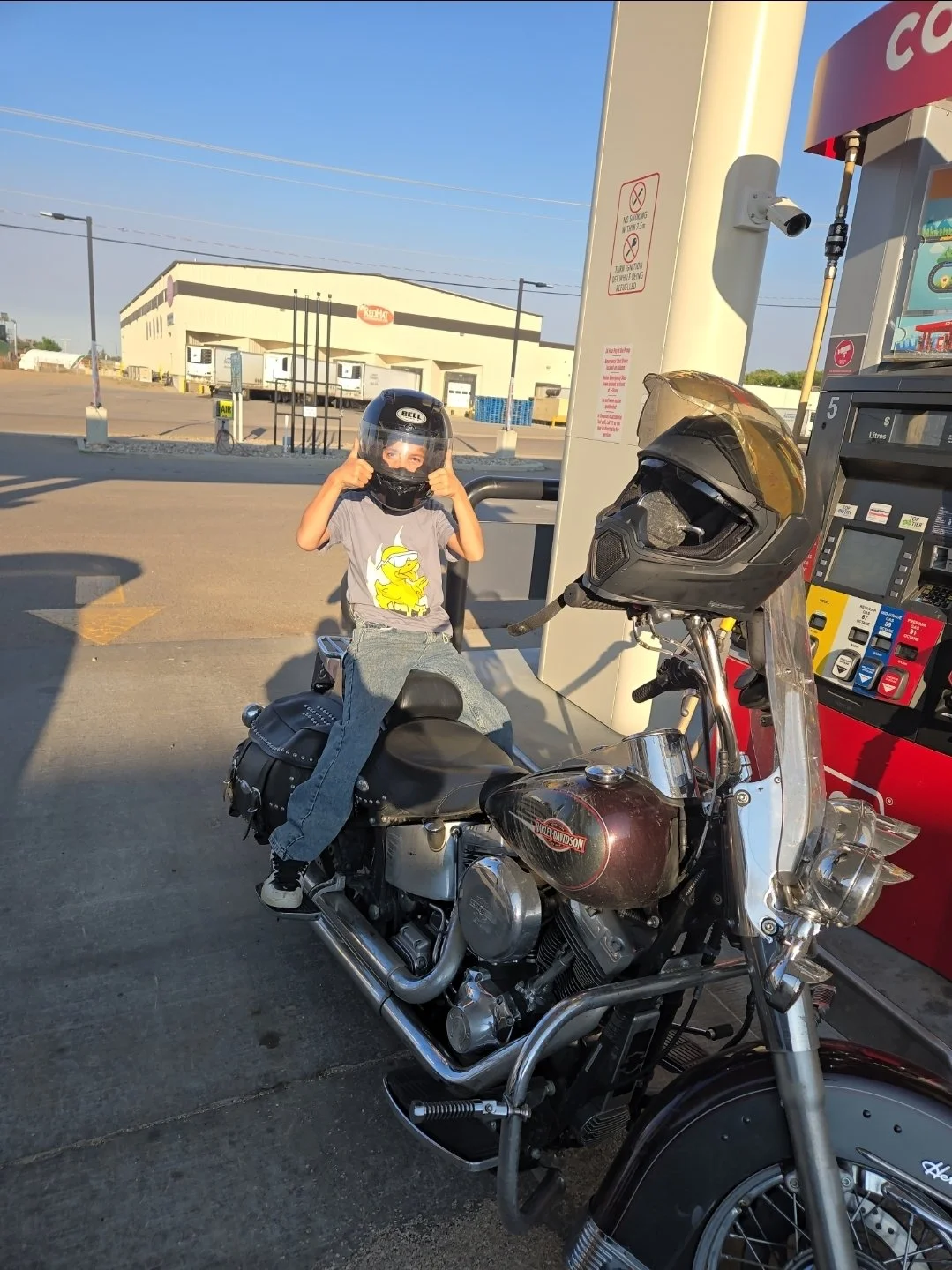 A young boy sitting on a Harley-Davidson motorcycle at a gas station, wearing a black helmet with a face shield and giving a thumbs-up, with a gray t-shirt featuring a yellow cartoon character.