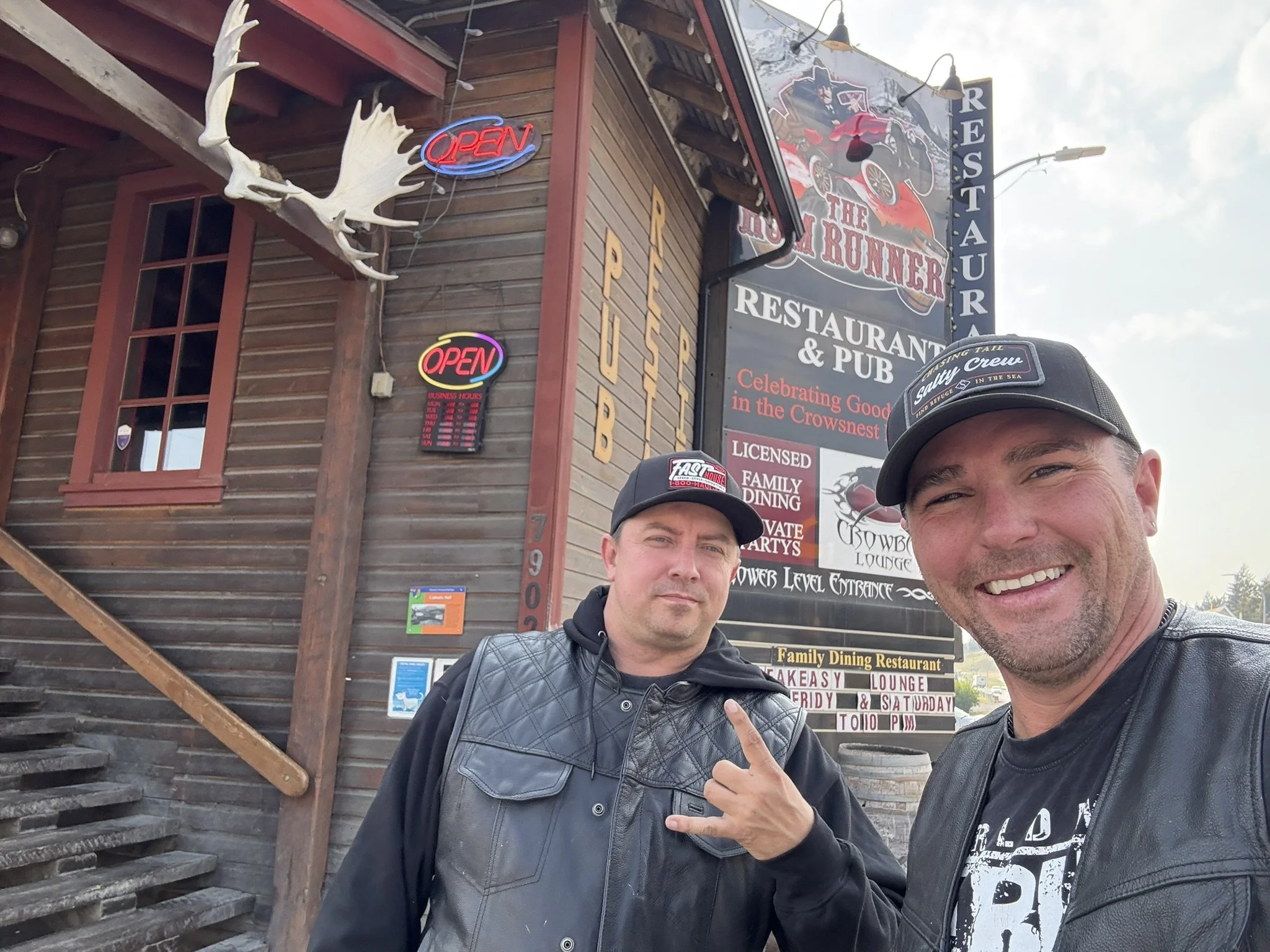Two men smiling in front of a restaurant sign, with one pointing at the sign. The restaurant is called The Rower, offering food, a pub, and family dining. The sign has a vintage car image and mentions celebrating good times. The building is wooden wi