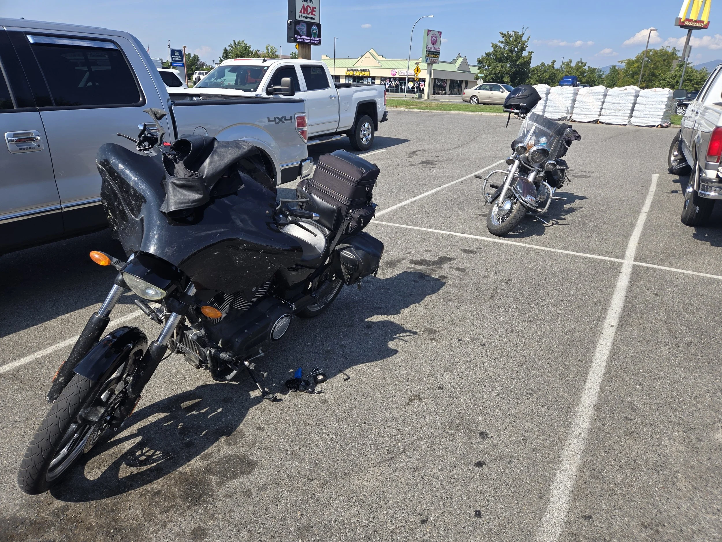 Two motorcycles parked in a parking lot near a silver pickup truck and other vehicles, with retail stores and a McDonald's visible in the background on a sunny day.