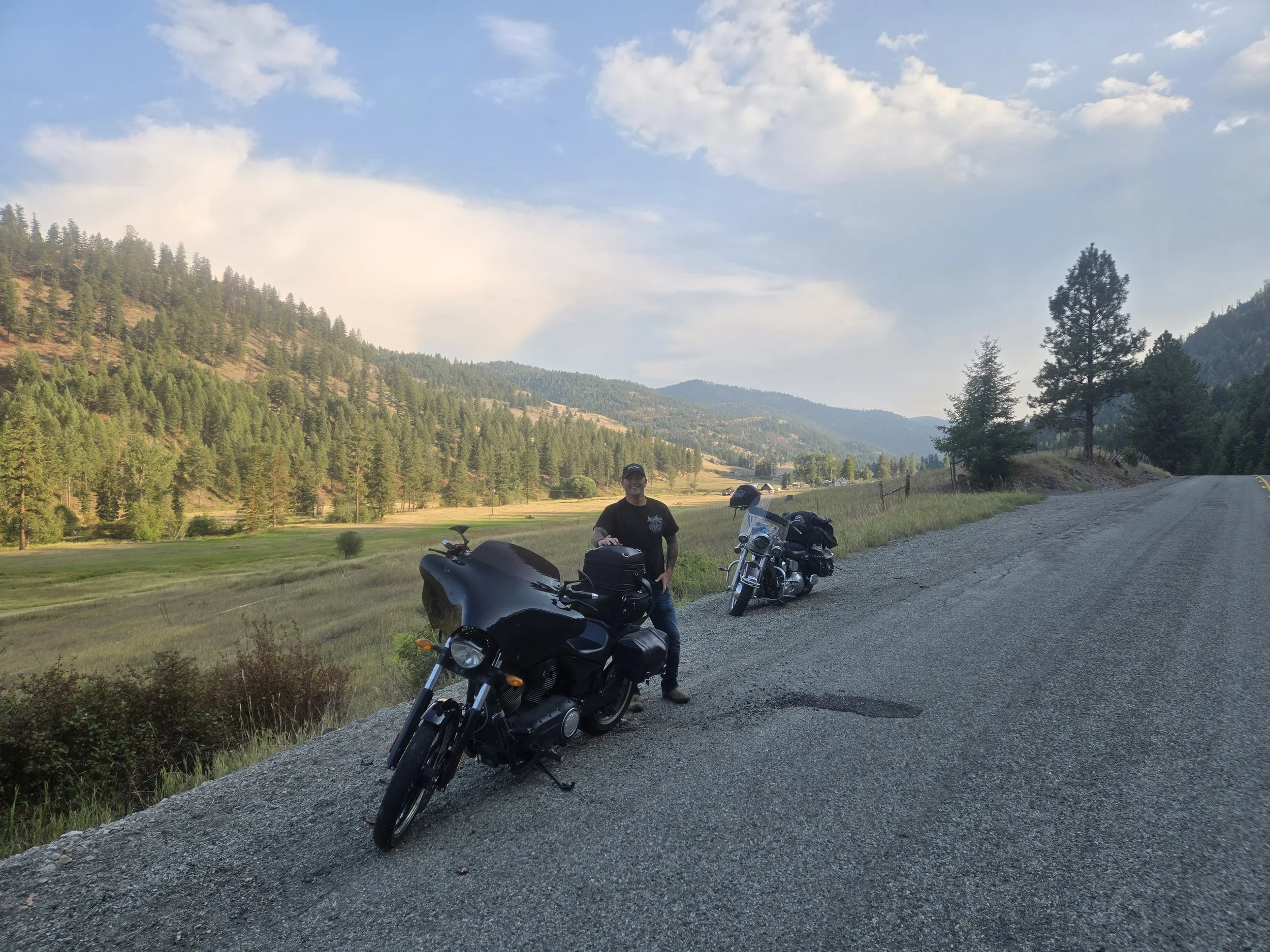 A man standing next to two motorcycles on a scenic mountain road with trees, hills, and a partly cloudy sky in the background.