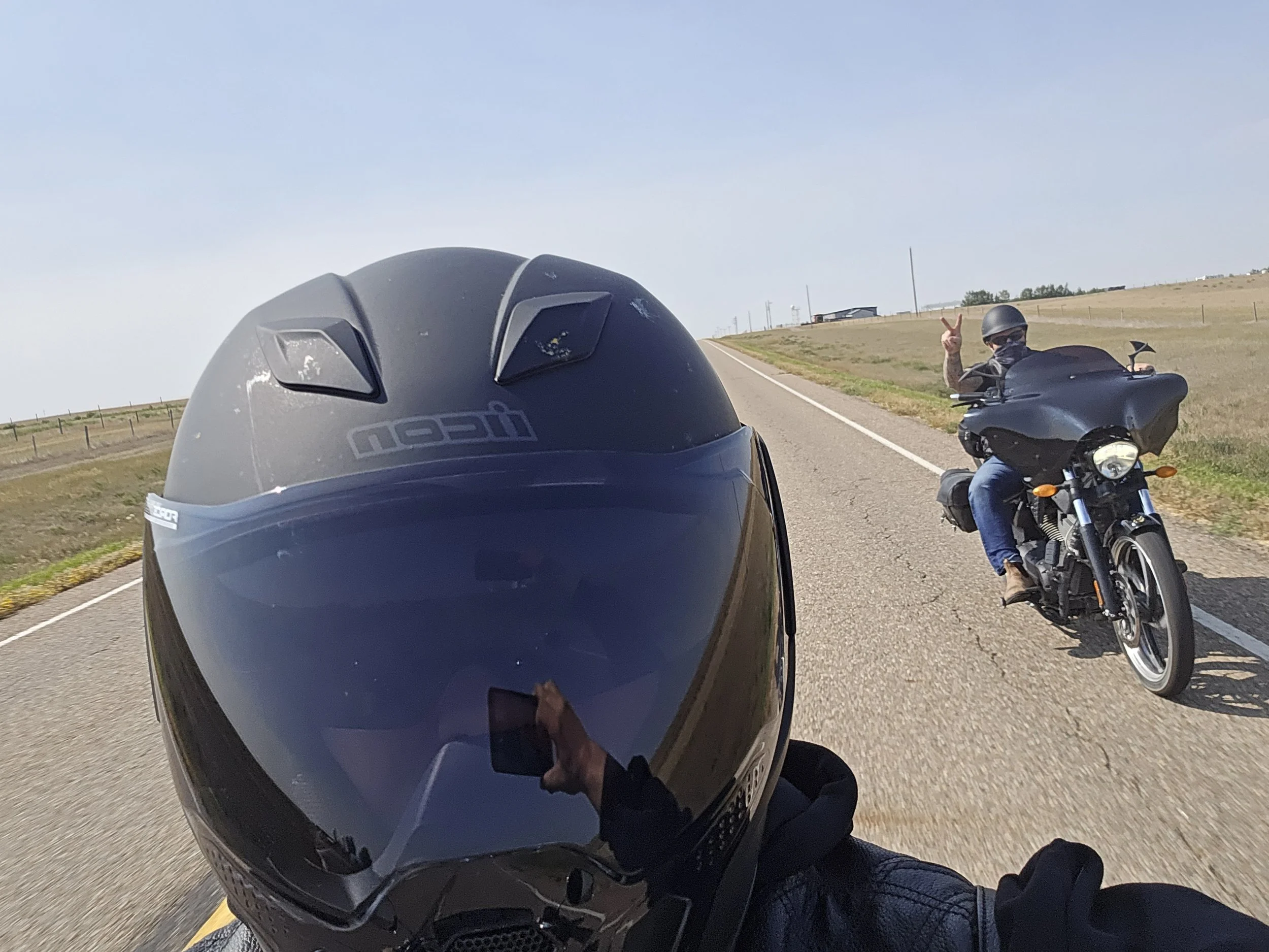 Motorcycle helmet in foreground, rider taking photo of a person on a motorcycle in the distance making a peace sign on an open road with grassy fields.