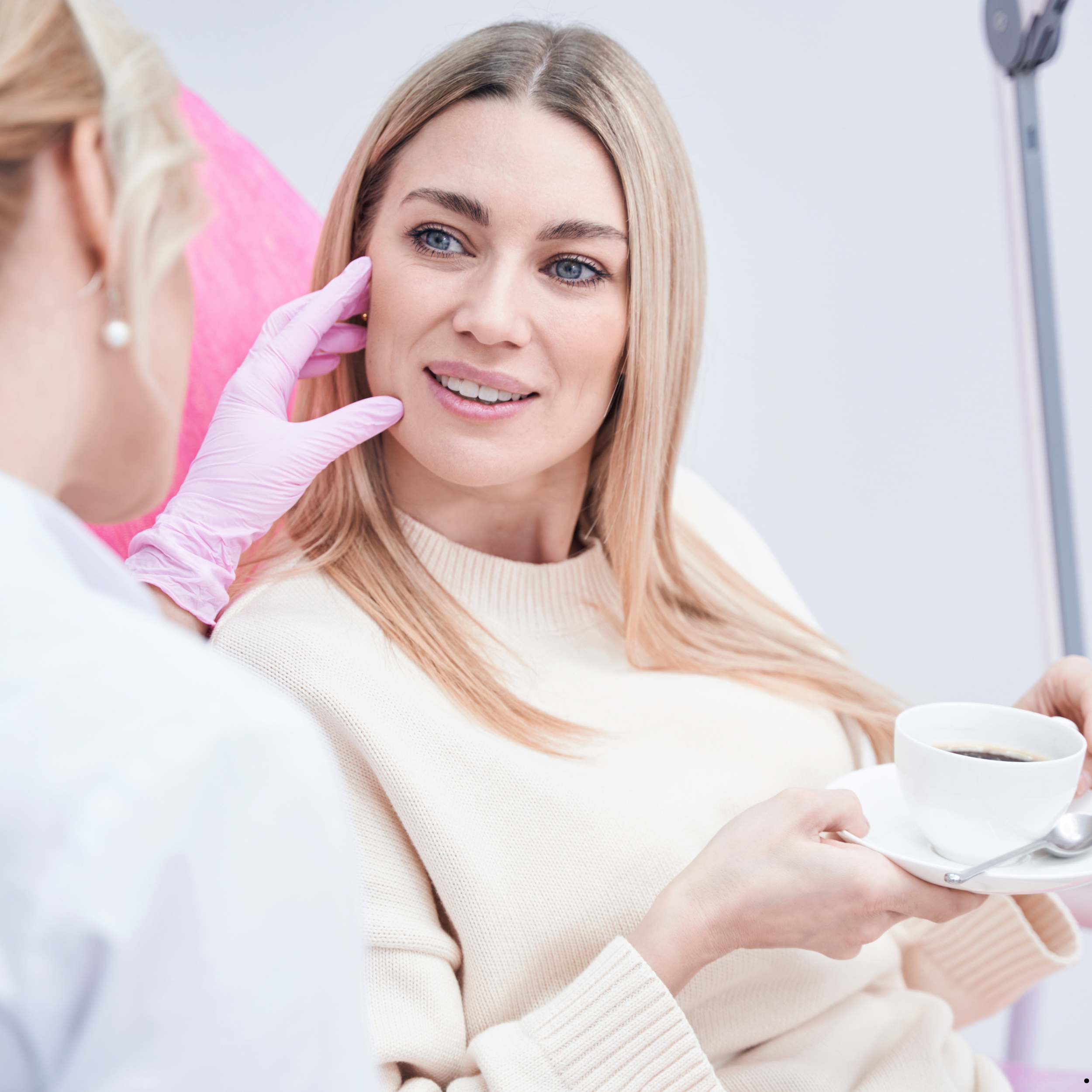 A woman with blonde hair and a light beige sweater is receiving a facial treatment from a professional wearing gloves. The woman is smiling and holding a white cup of coffee with a saucer and spoon.