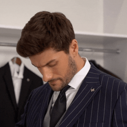 A man in a navy pinstripe suit with a white shirt and black tie looking down in a clothing store fitting room.