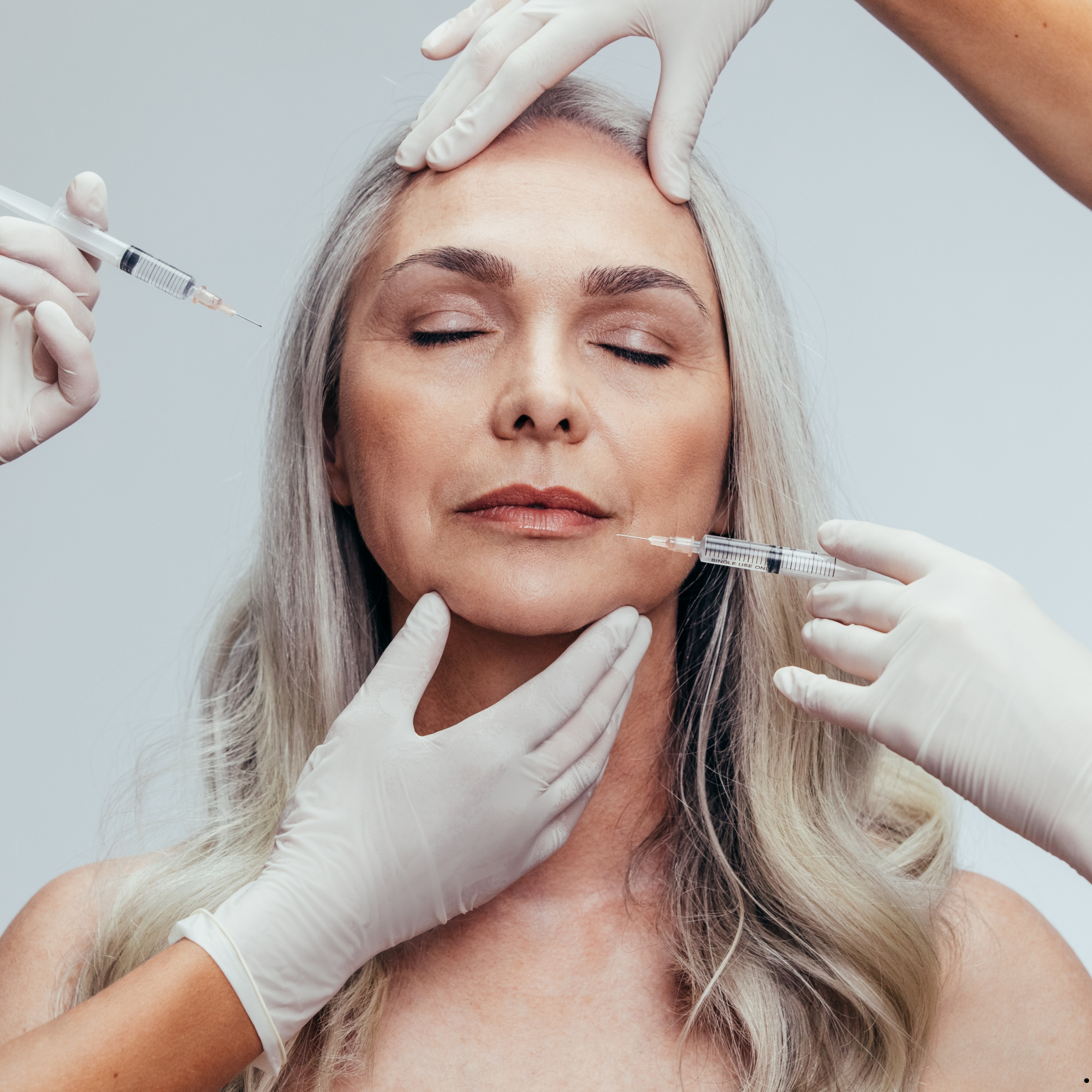 A woman receiving a cosmetic injection with closed eyes, surrounded by four gloved hands holding syringes, in a clinical setting.