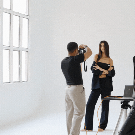 A woman posing for a photo taken by a man in a bright photo studio with white walls and large windows.