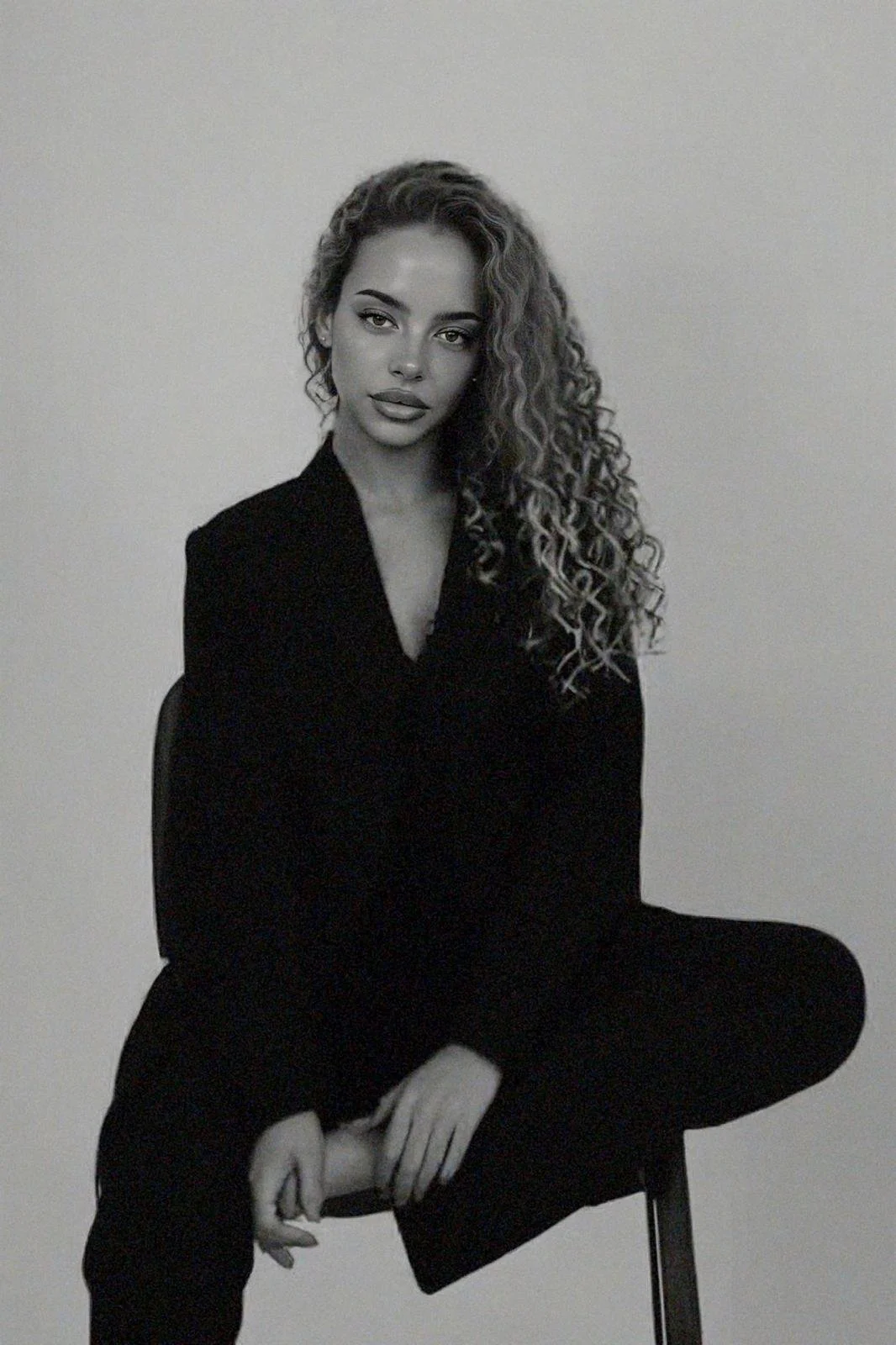 A black-and-white photo of a woman with long, curly hair sitting on a chair against a plain background. She is wearing a dark blazer and pants, looking directly at the camera with a neutral expression.