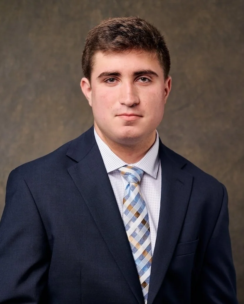 A young man in a dark suit, light-colored shirt, and patterned tie posing for a formal portrait against a plain brown background.