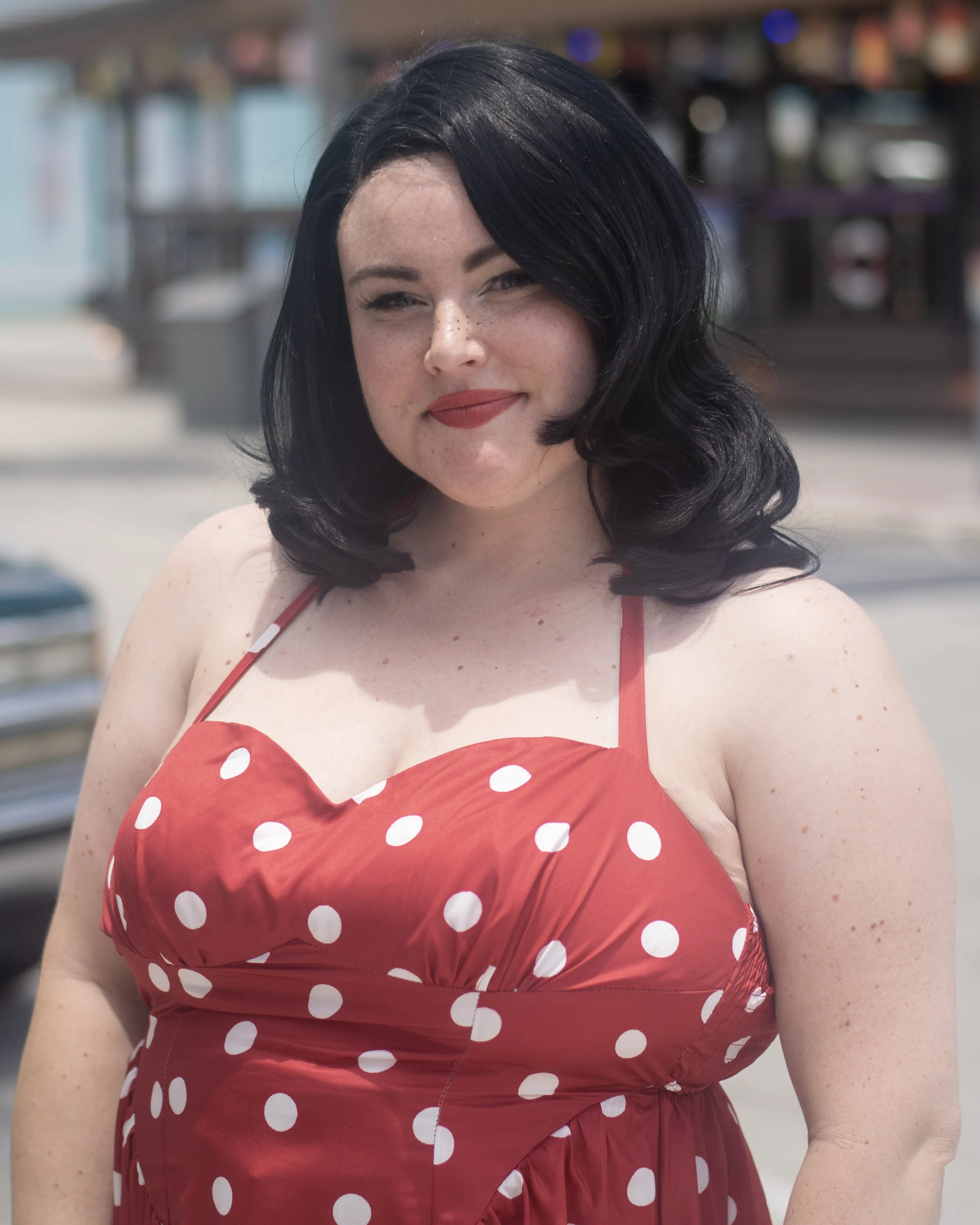A woman with fair skin, dark hair, and freckles wearing a red polka dot dress with spaghetti straps at an outdoor location.