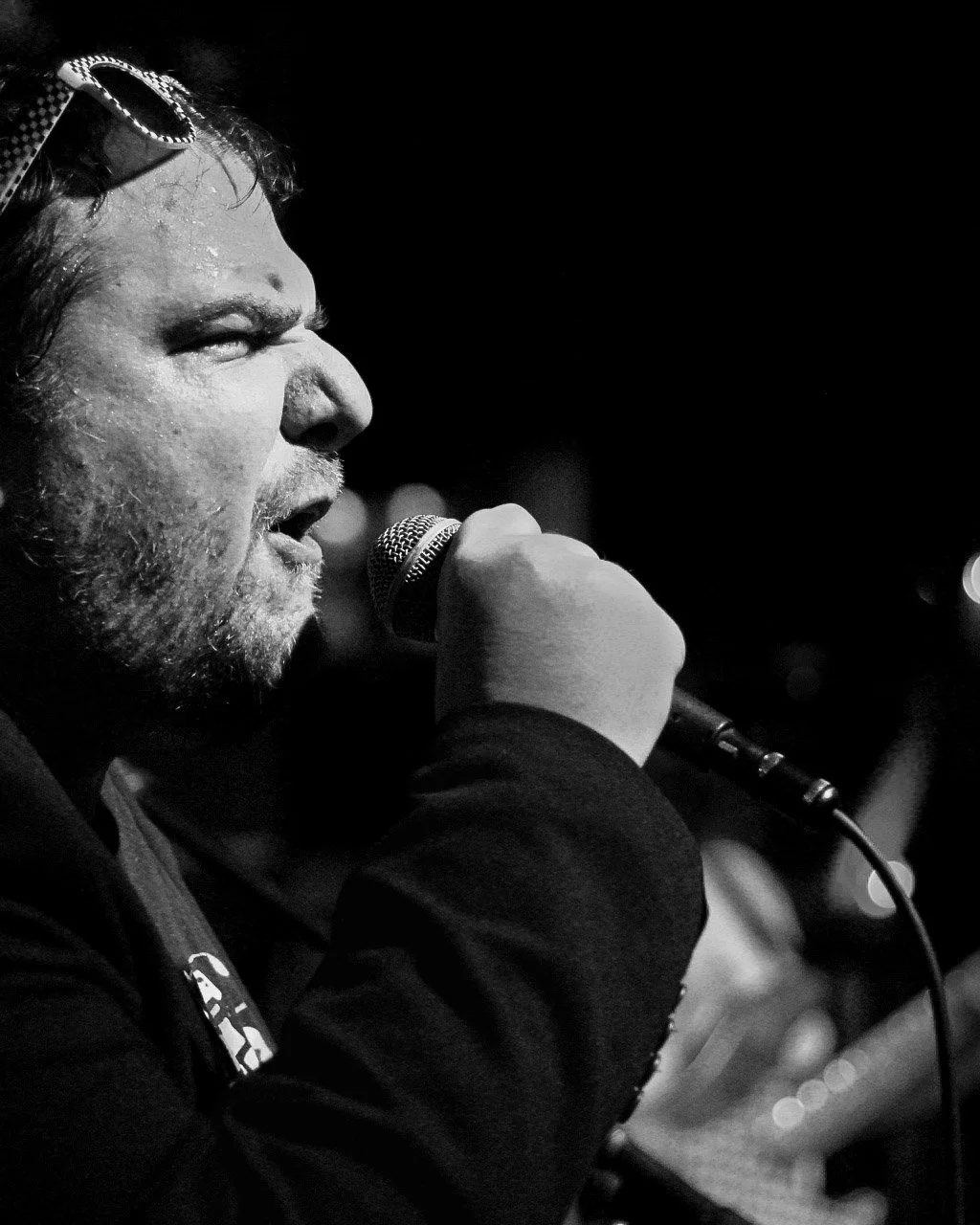 A black and white photograph of a man with glasses pushed up on his head, singing into a microphone, with a dark blurred background.