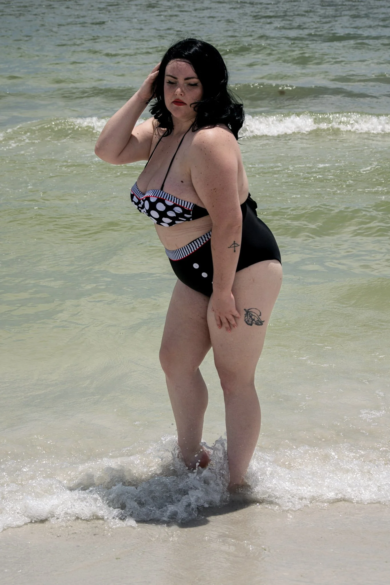 A woman with black hair, wearing a polka dot swimsuit, stands in shallow water at the beach, with waves and sand visible in the background.