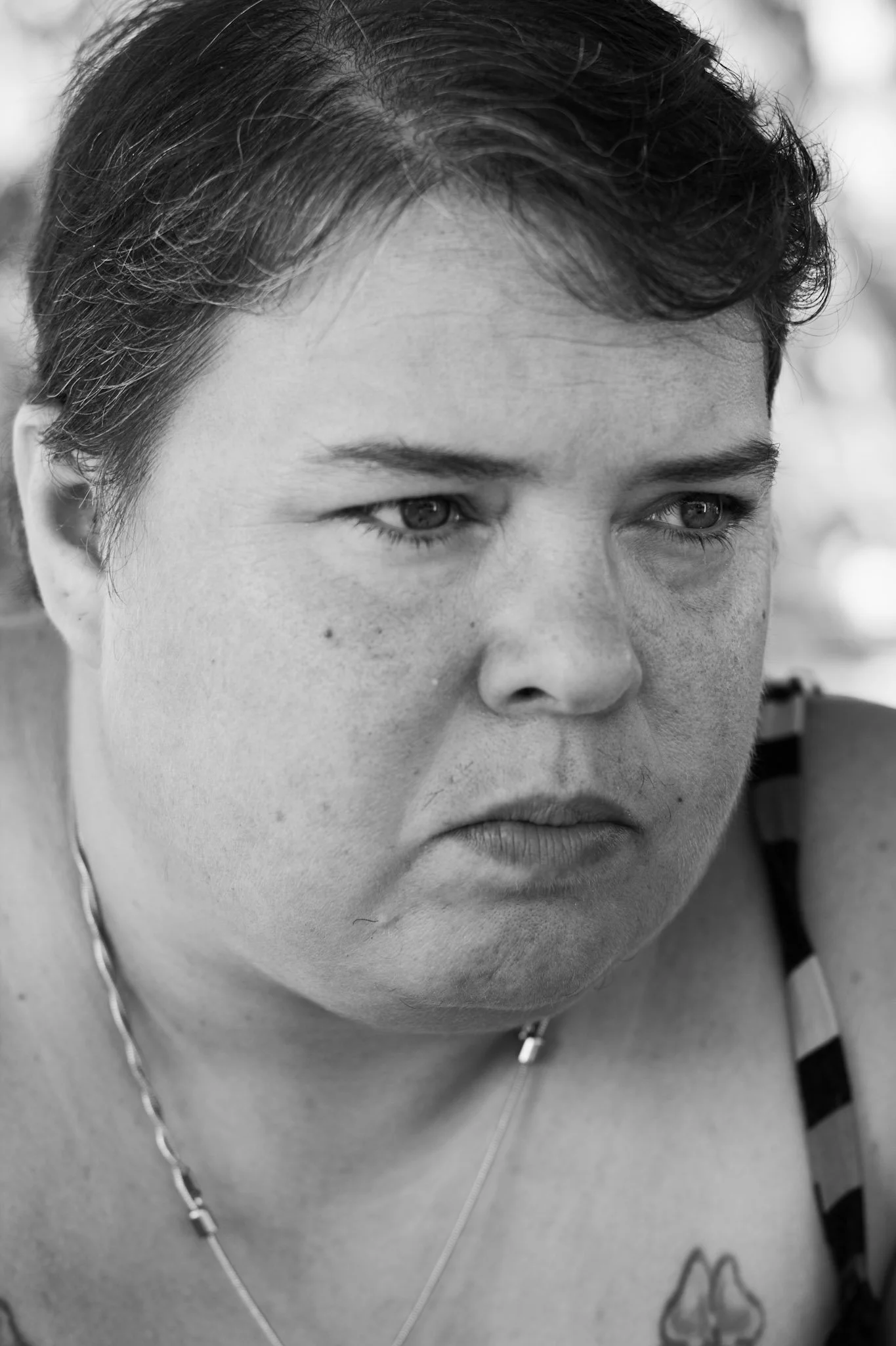 Close-up of a woman with short dark hair, looking downward, with a serious expression, wearing a necklace and a shirt with a striped pattern.
