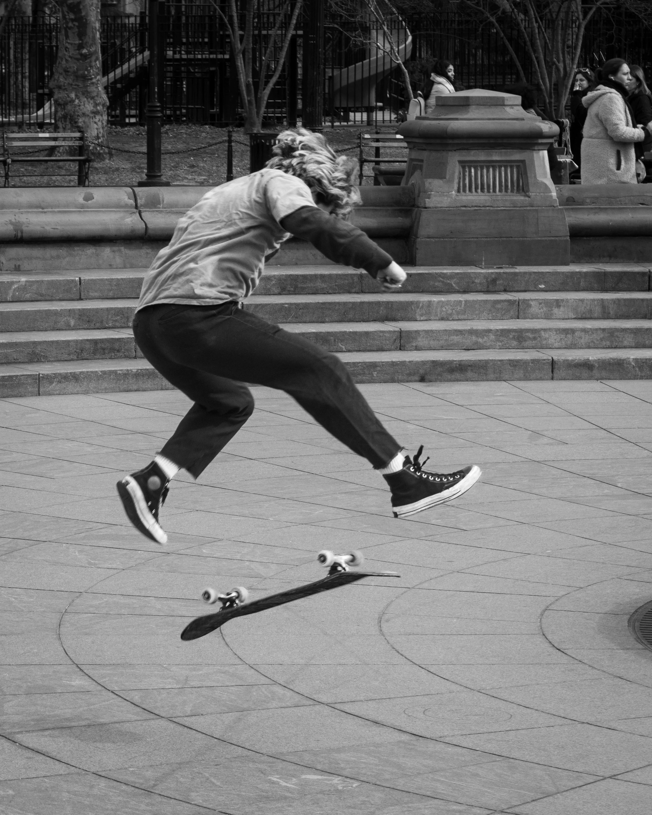 A person with long hair, wearing a t-shirt and jeans, is doing a kickflip on a skateboard on steps in an urban park, with several people standing in the background near benches and trees.