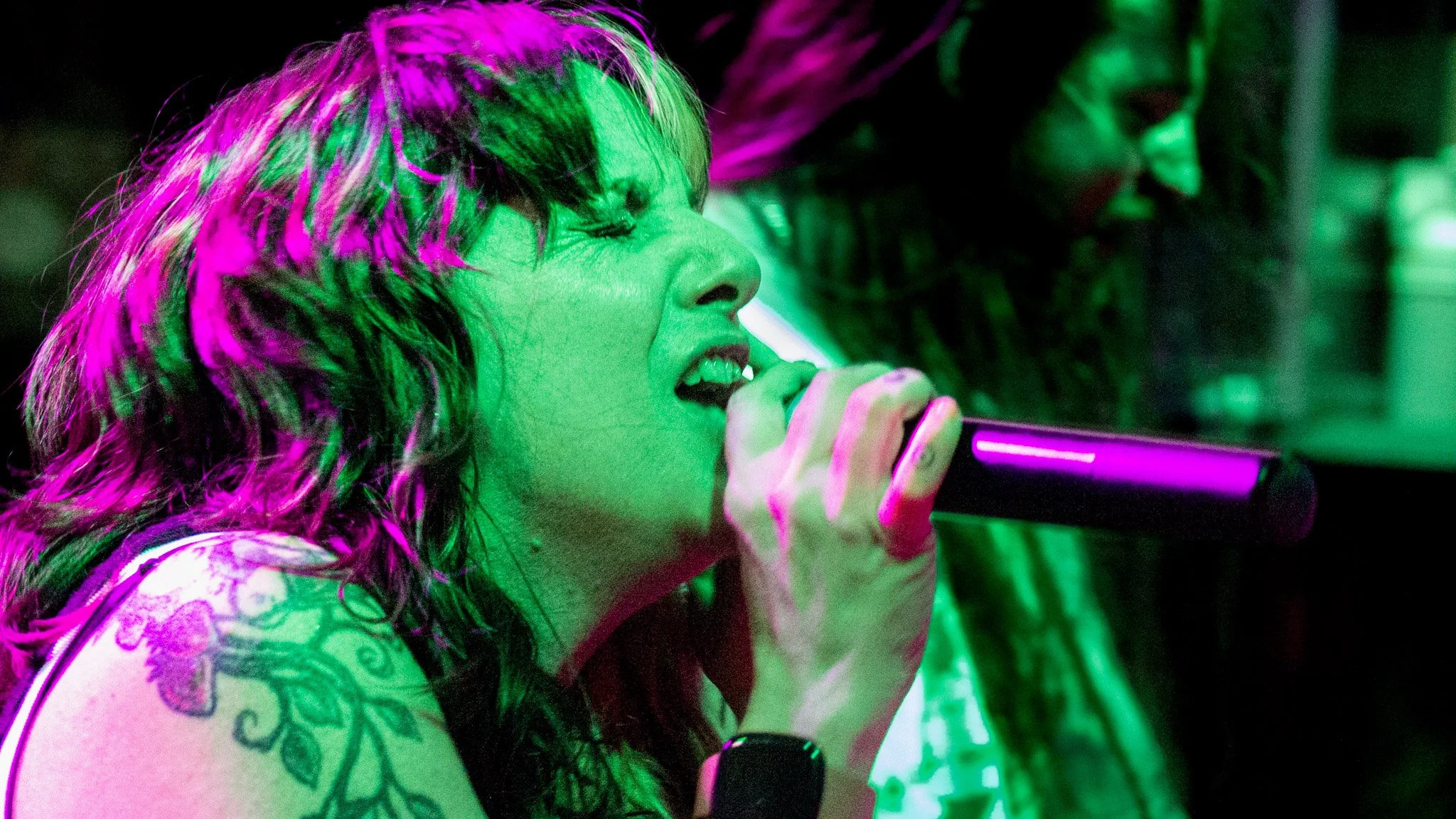 A woman with multicolored hair singing into a microphone, illuminated by vibrant green and pink lighting, with tattoos visible on her arm.