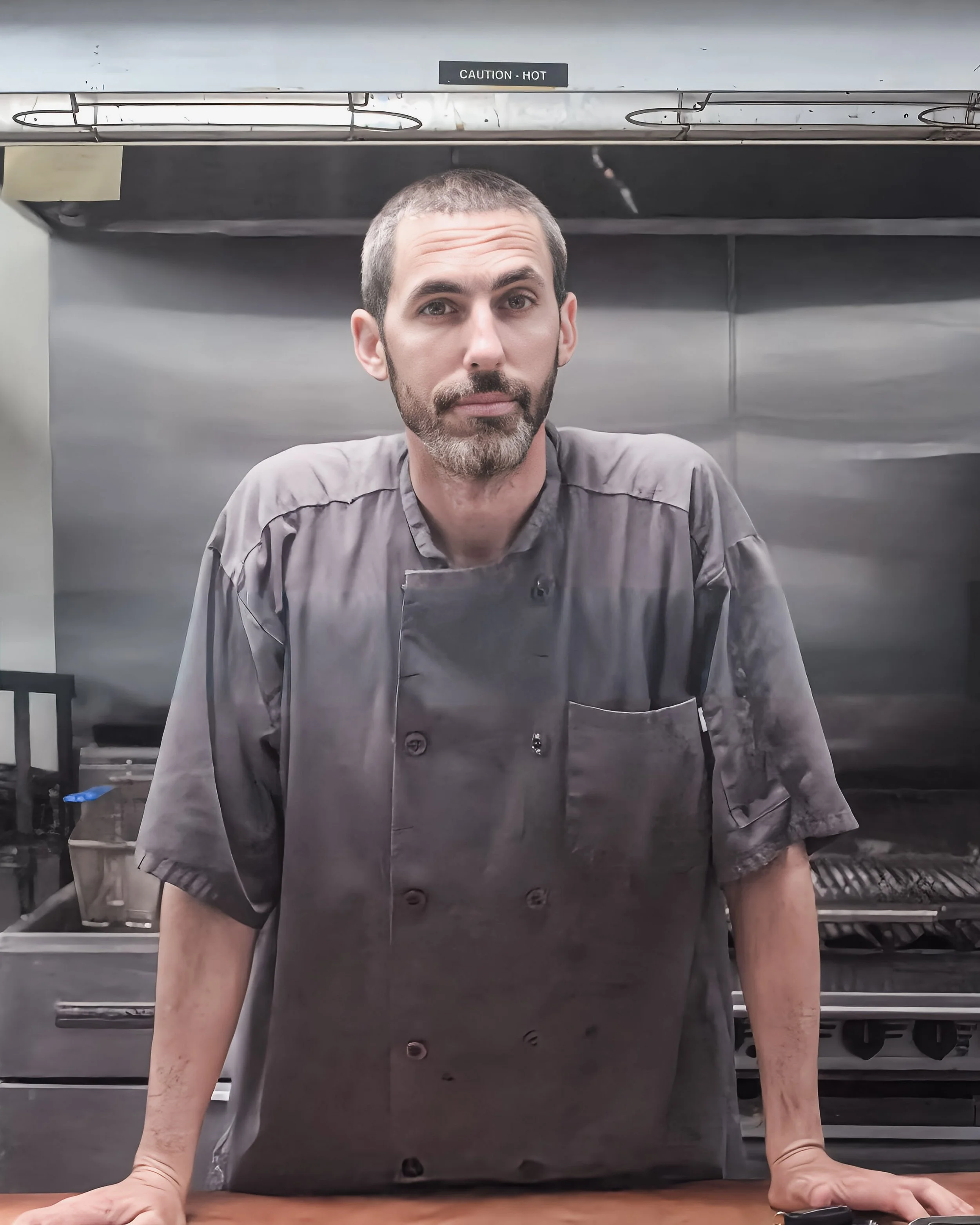 A man with a beard and short hair in a gray chef's coat is standing in a commercial kitchen, with his hands resting on a wooden surface. There is a large stainless steel oven or stove behind him, with a caution sign that reads 'CAUTION - HOT' on top.