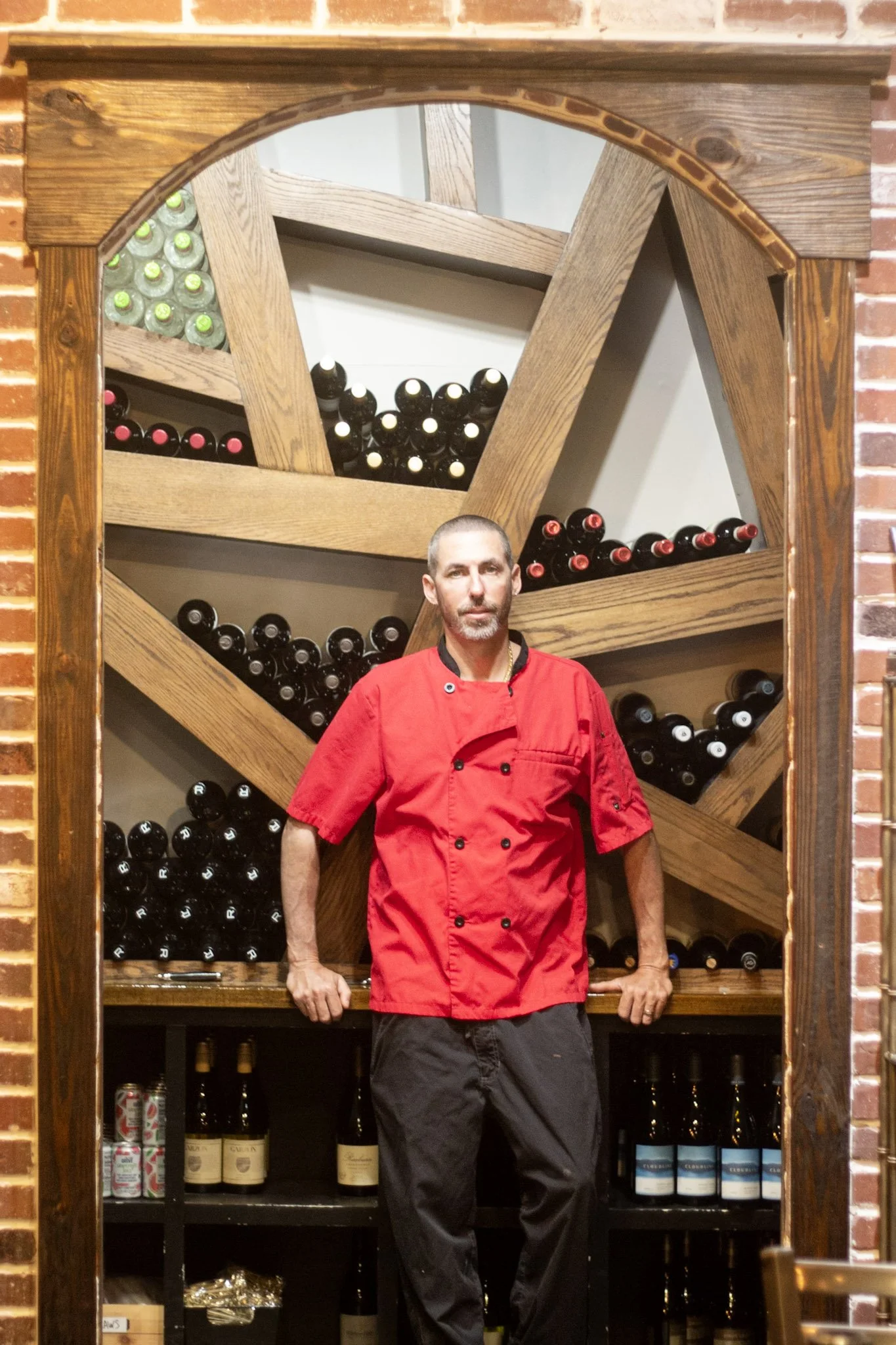 A man in a red chef jacket standing in front of wine bottles on a wine rack, with a decorative wooden and brick arch behind him.