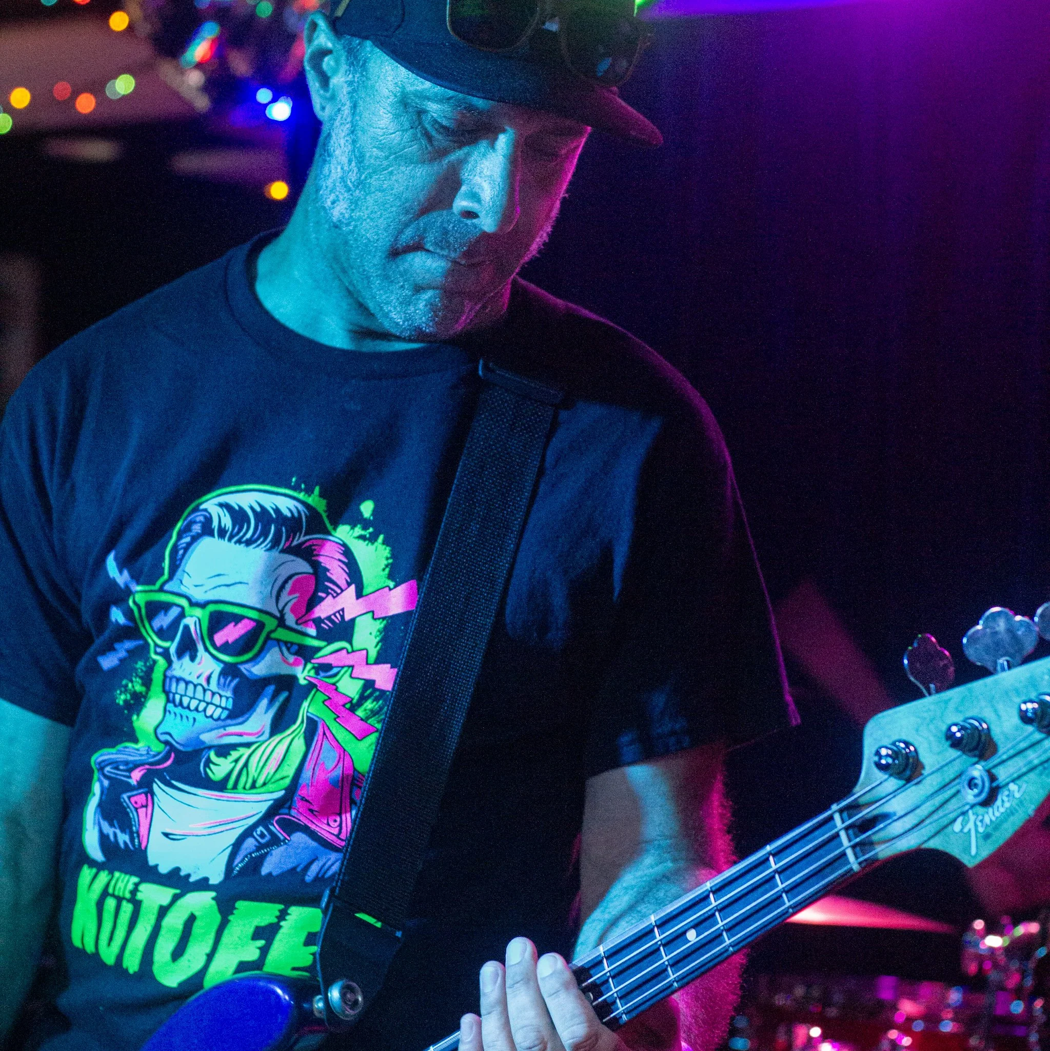 A person wearing glasses and a hat is playing an electric guitar in a dimly lit environment with multicolored lights. The individual is dressed in a black shirt with a colorful graphic design.