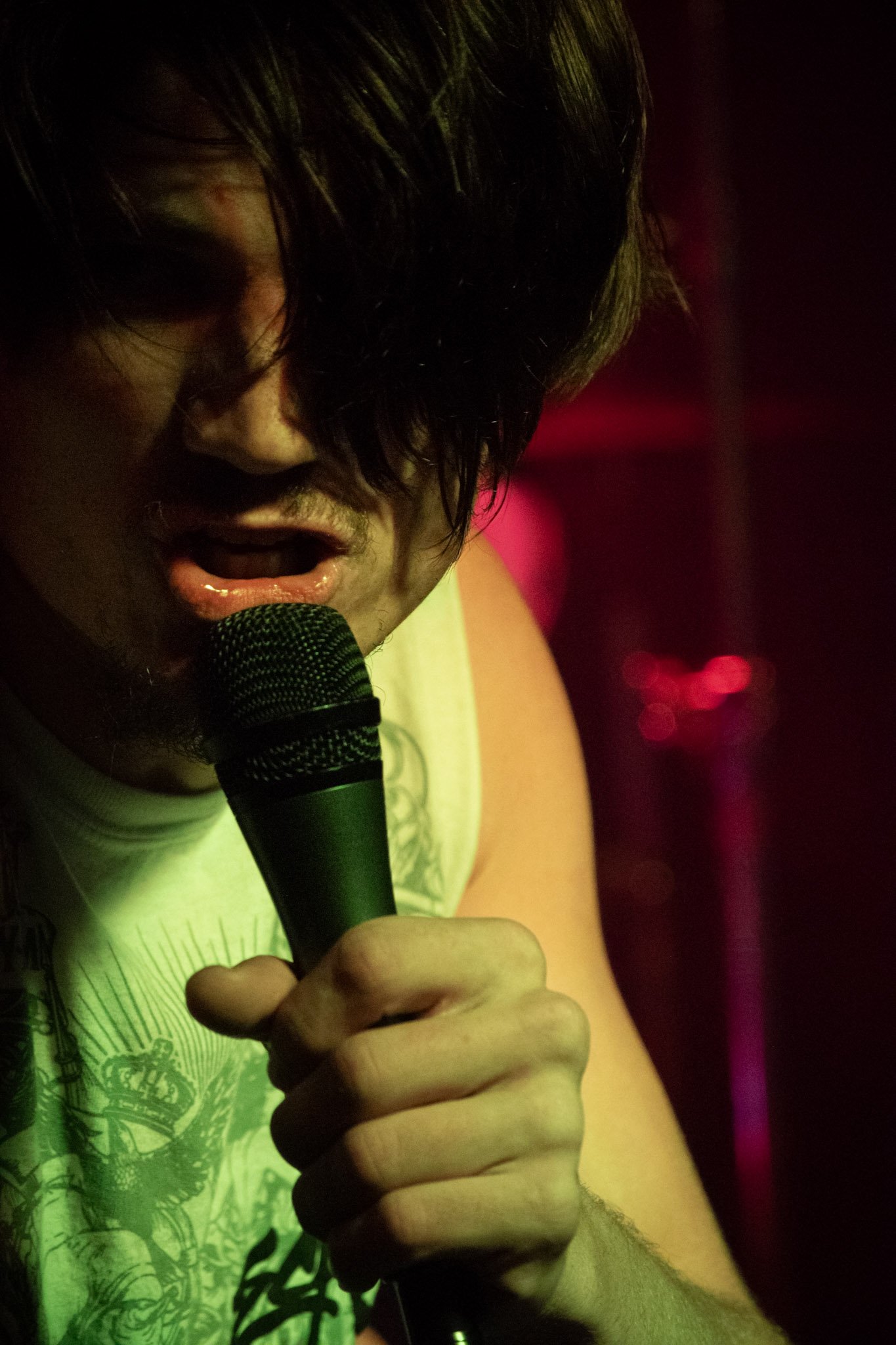 A person with dark hair and holding a microphone close to their mouth and singing, with colorful concert lighting in the background.