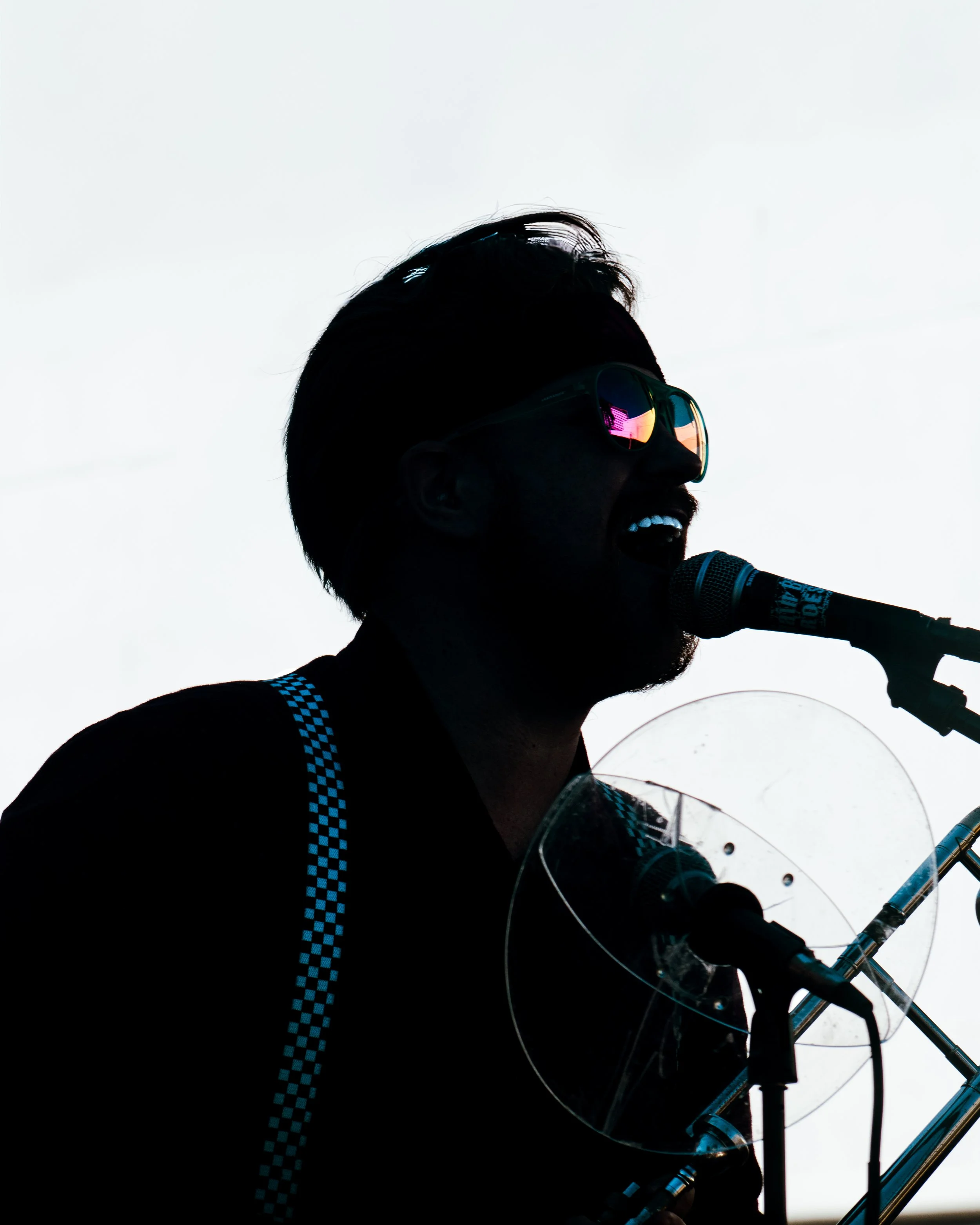 A person with a trombone, wearing sunglasses, singing into a microphone, in silhouette.