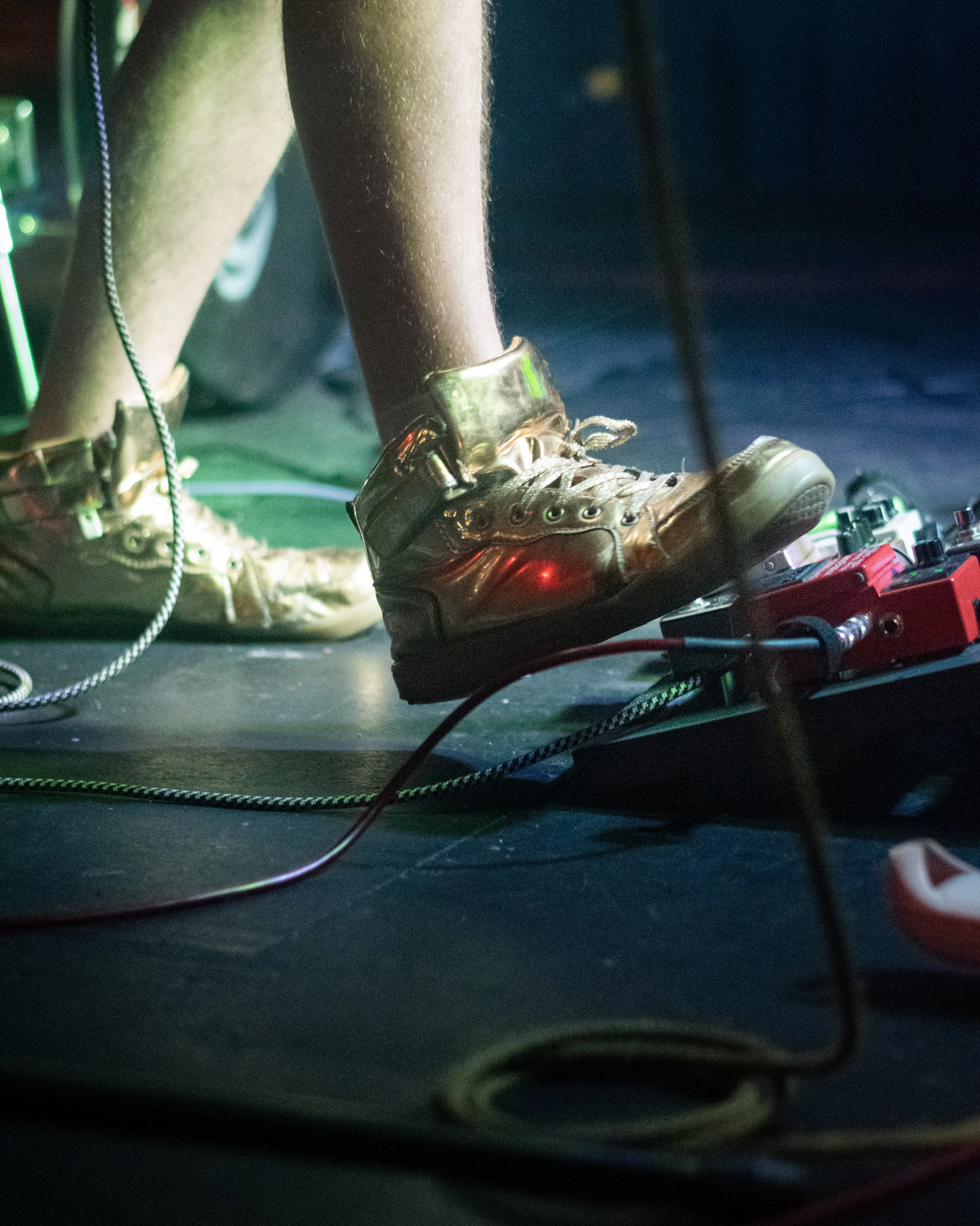 Close-up of a person's leg wearing gold-colored high-top sneakers, standing on a guitar pedal with various cables and equipment around in a dimly lit setting with green and red lighting.