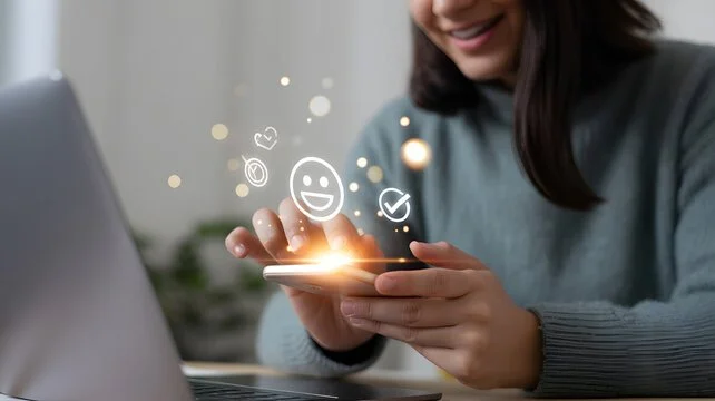 A woman smiling while using a smartphone with digital icons and emojis floating above it.