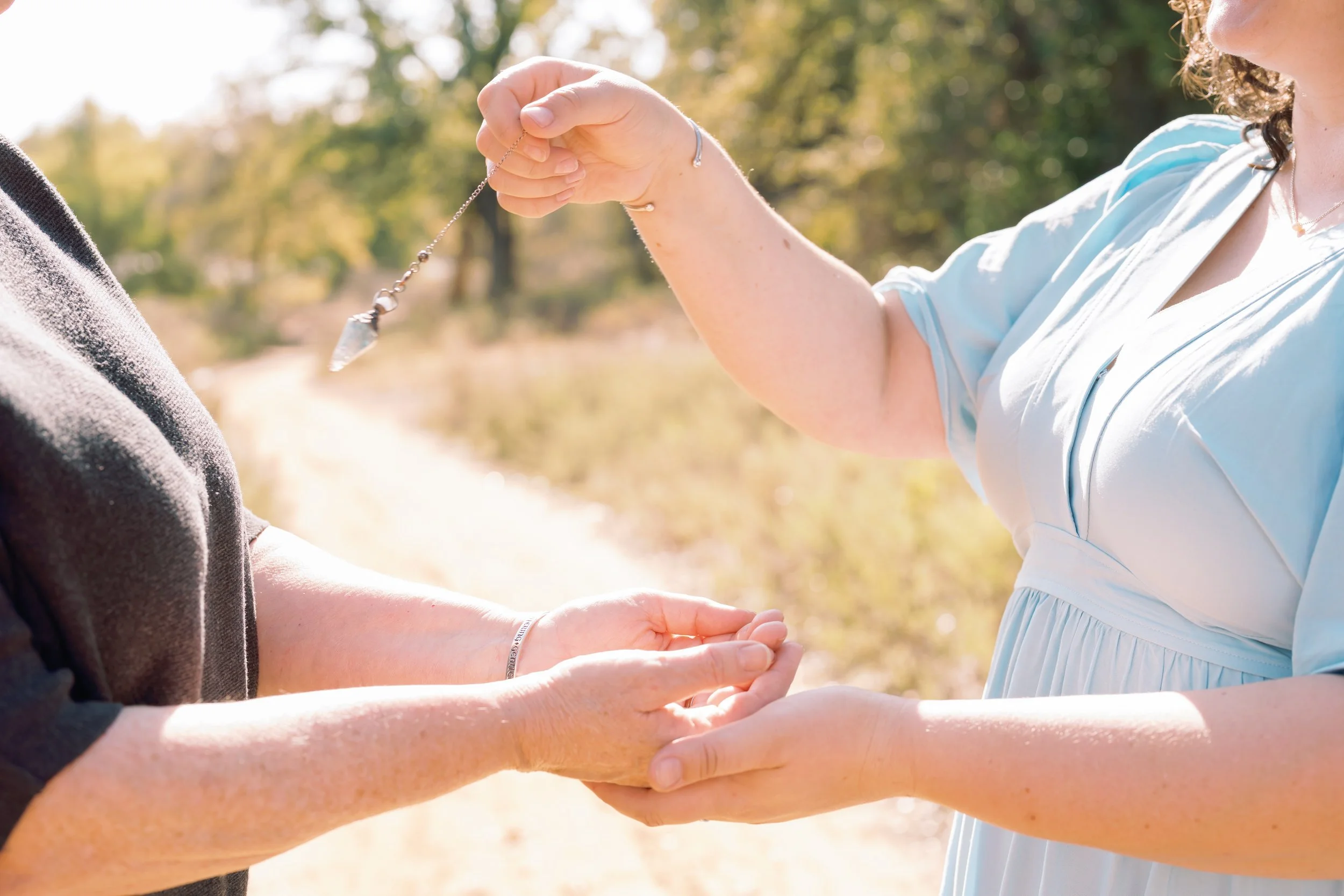 A woman in a light blue dress is placing a necklace around another person's neck during an outdoor ceremony. The scene is bathed in sunlight with trees and a pathway in the background.