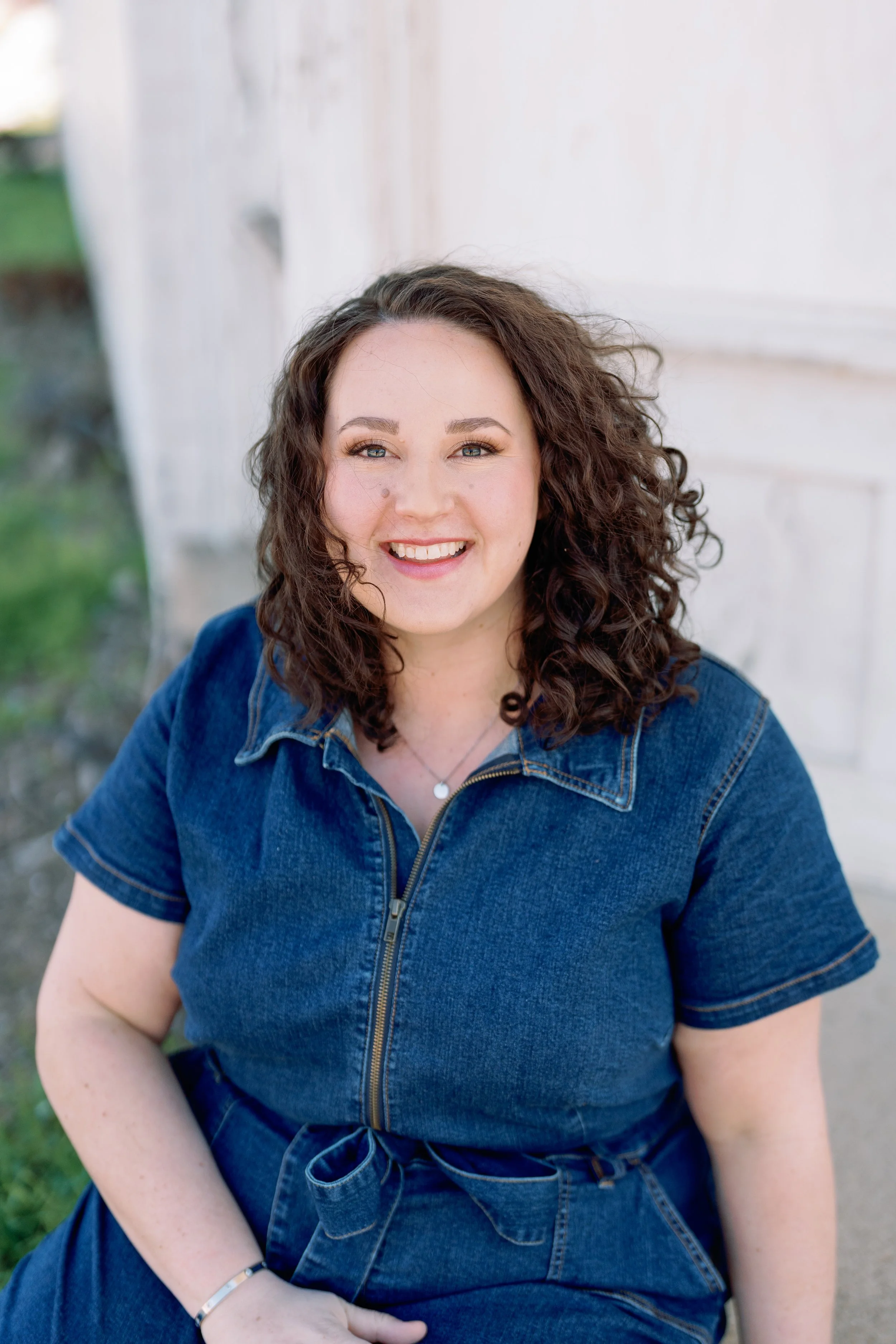 Portrait of a smiling woman with curly brown hair, wearing a denim jumpsuit, seated outdoors against a white wall.