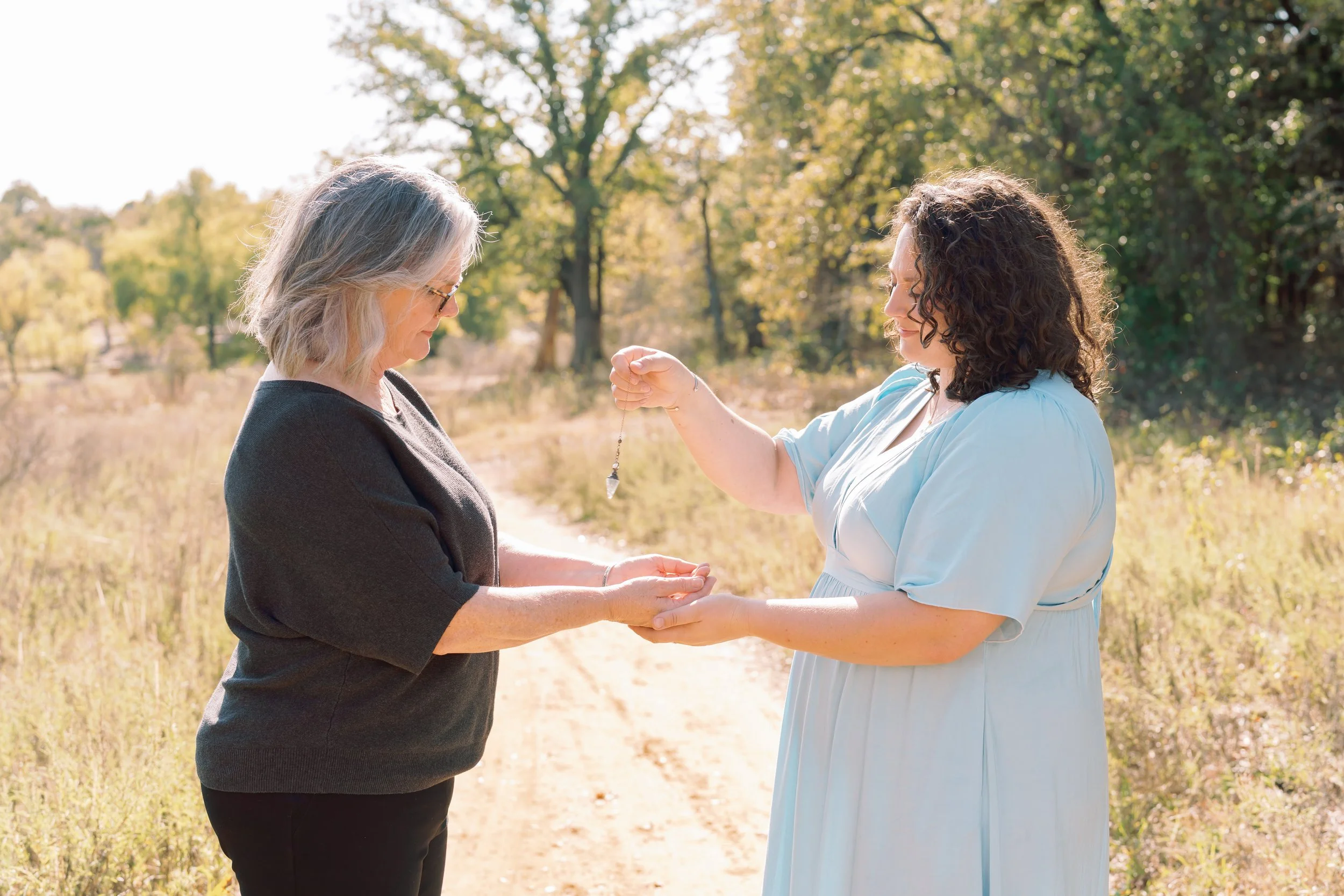 Two women exchanging jewelry outside on a sunny day, one woman gives a necklace to another, with trees and a dirt path in the background.
