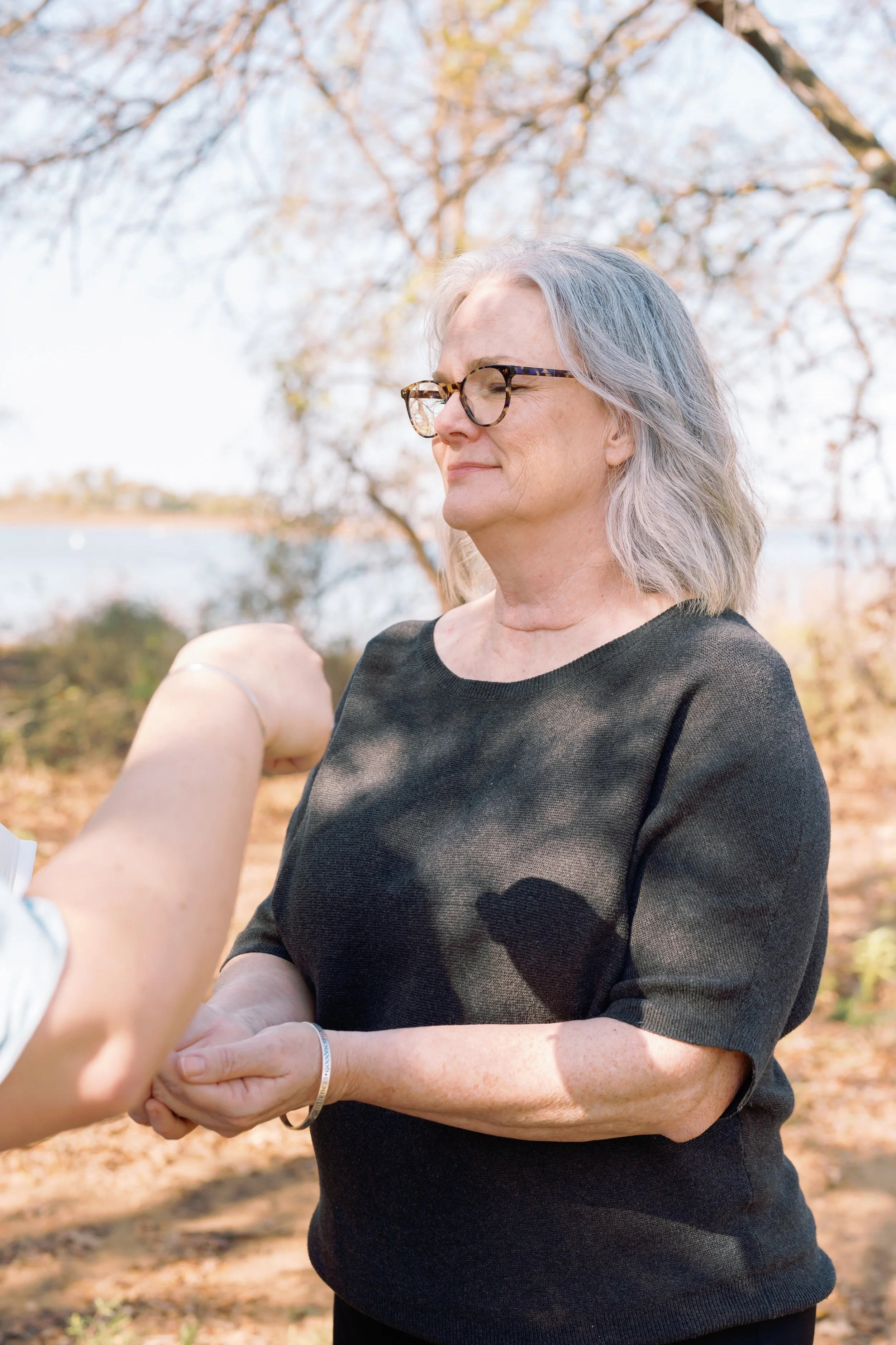 Older woman with gray hair and glasses receives a ring in an outdoor setting with trees and a lake in the background.