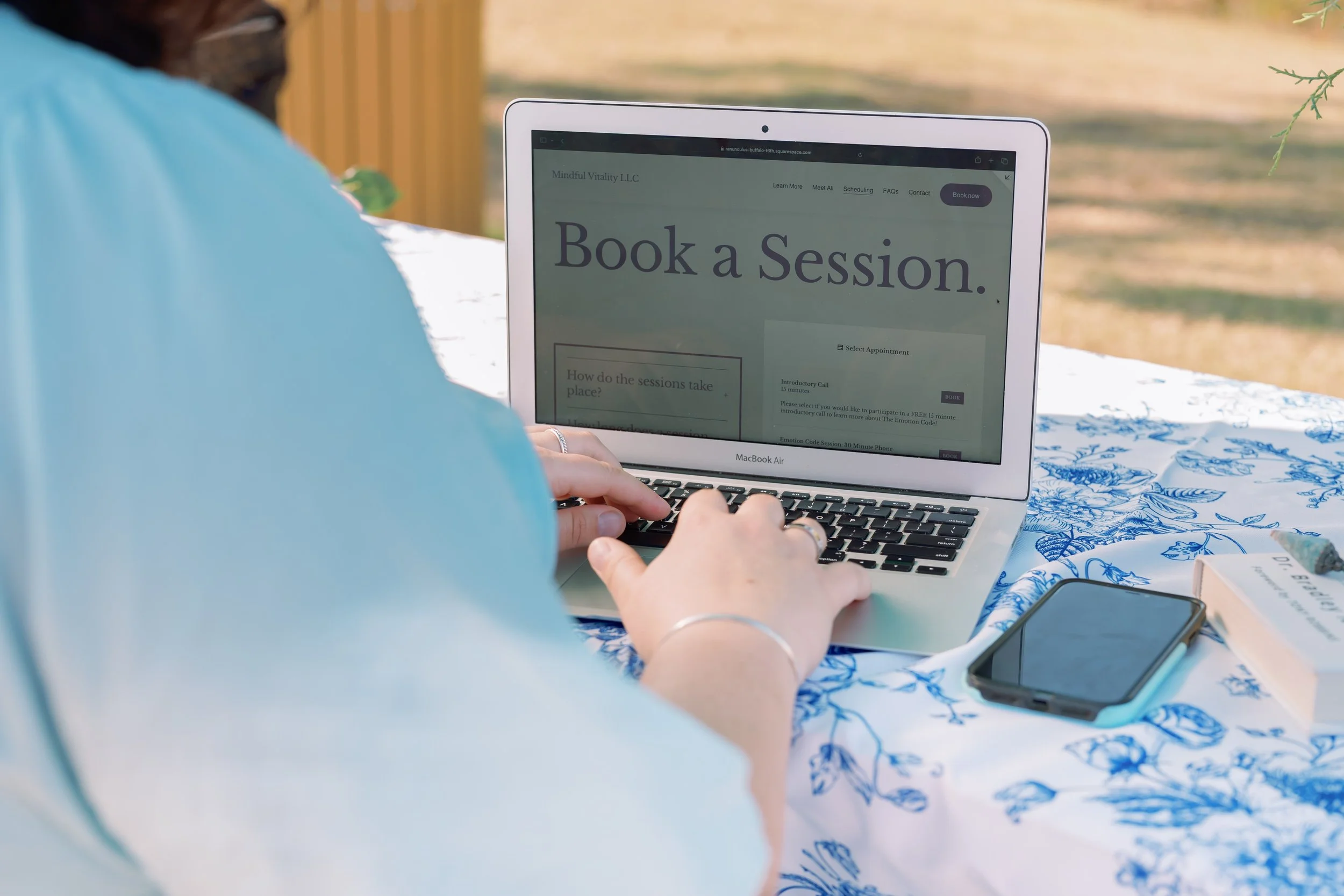 Person using a laptop outdoors with a smartphone and a book on a table, viewing a webpage with 'Book a Session' text.