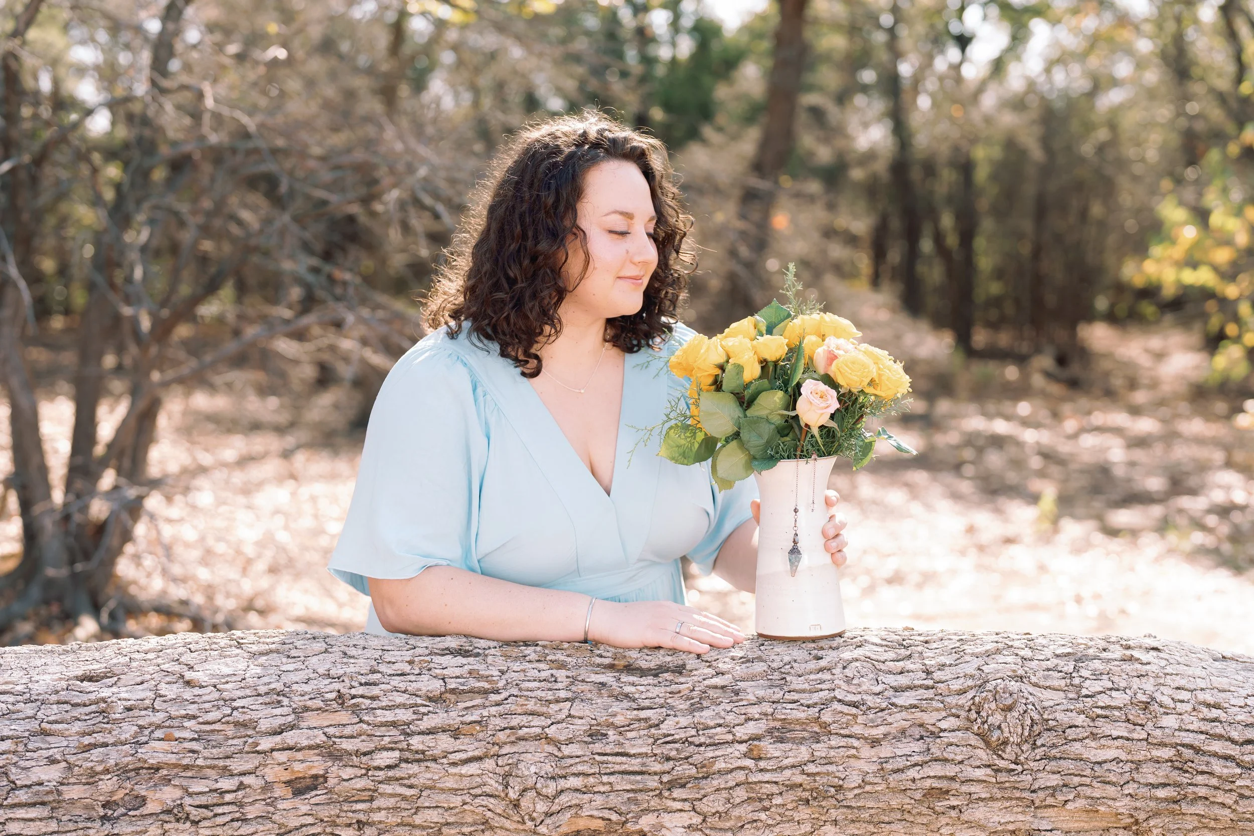 A woman with curly hair in a light blue dress holding a yellow and pink flower bouquet in a white vase outdoors during daytime.