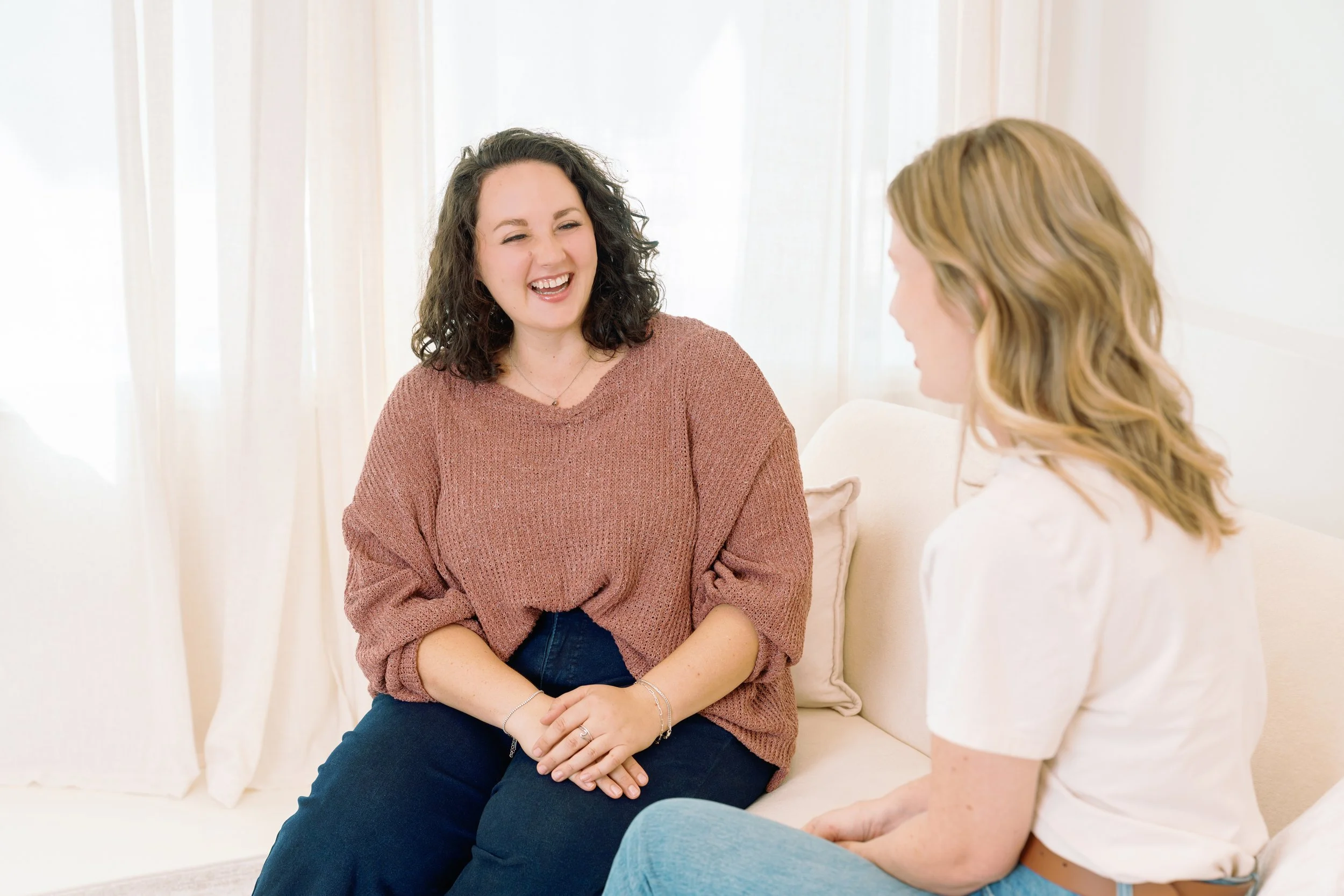 Two women are sitting on a white couch in a bright room with sheer curtains. They are engaged in a friendly conversation, smiling and making eye contact. The woman on the left has dark curly hair and is wearing a rust-colored sweater, while the woman on the right has long blonde hair and is wearing a white top.
