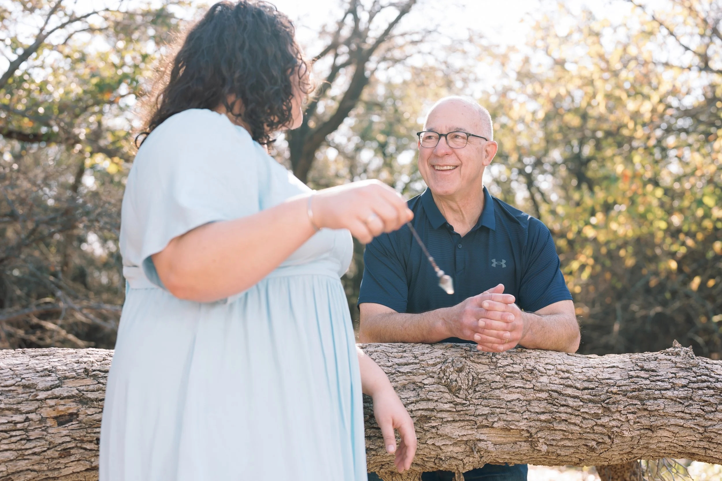 A woman in a light blue dress holding a necklace in front of an older man in a dark blue polo shirt, who is smiling and standing behind a fallen tree trunk.