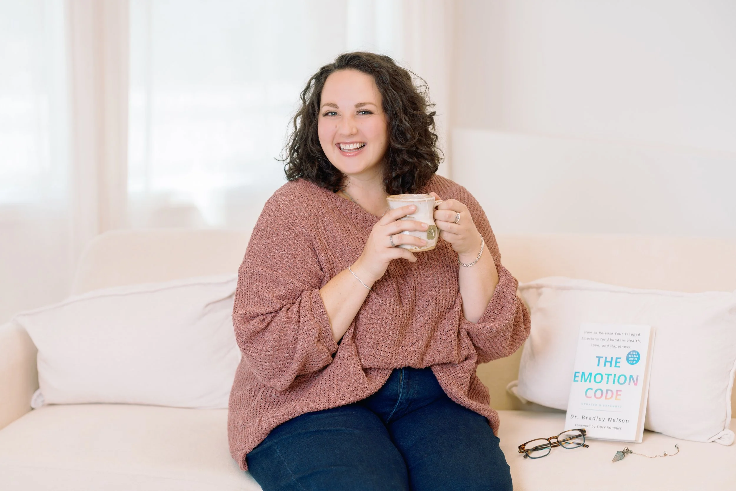 A woman with curly dark hair sitting on a white couch, smiling and holding a mug. There is a book titled "The Emotion Code" on the couch next to her.