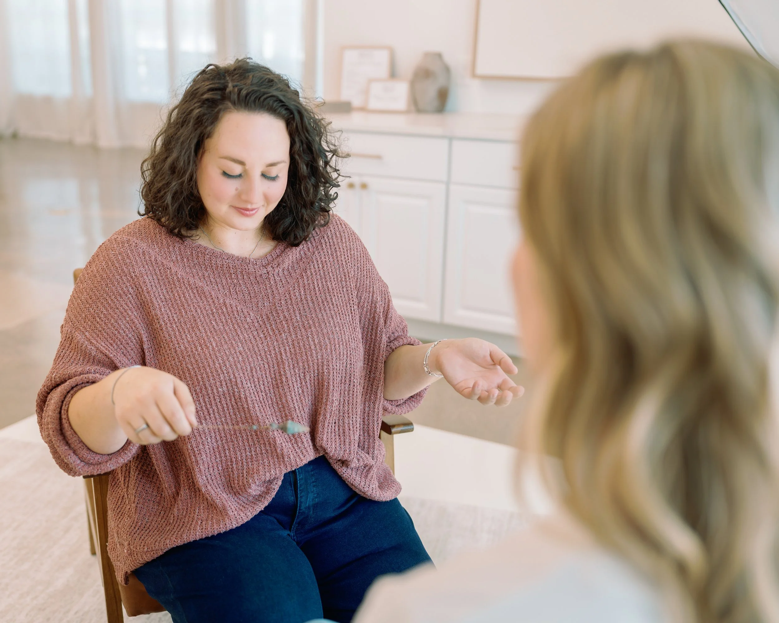 A woman with dark curly hair wearing a pink sweater sitting on a chair in a room with white furniture, engaged in a craft activity, with another person partially visible in the foreground.