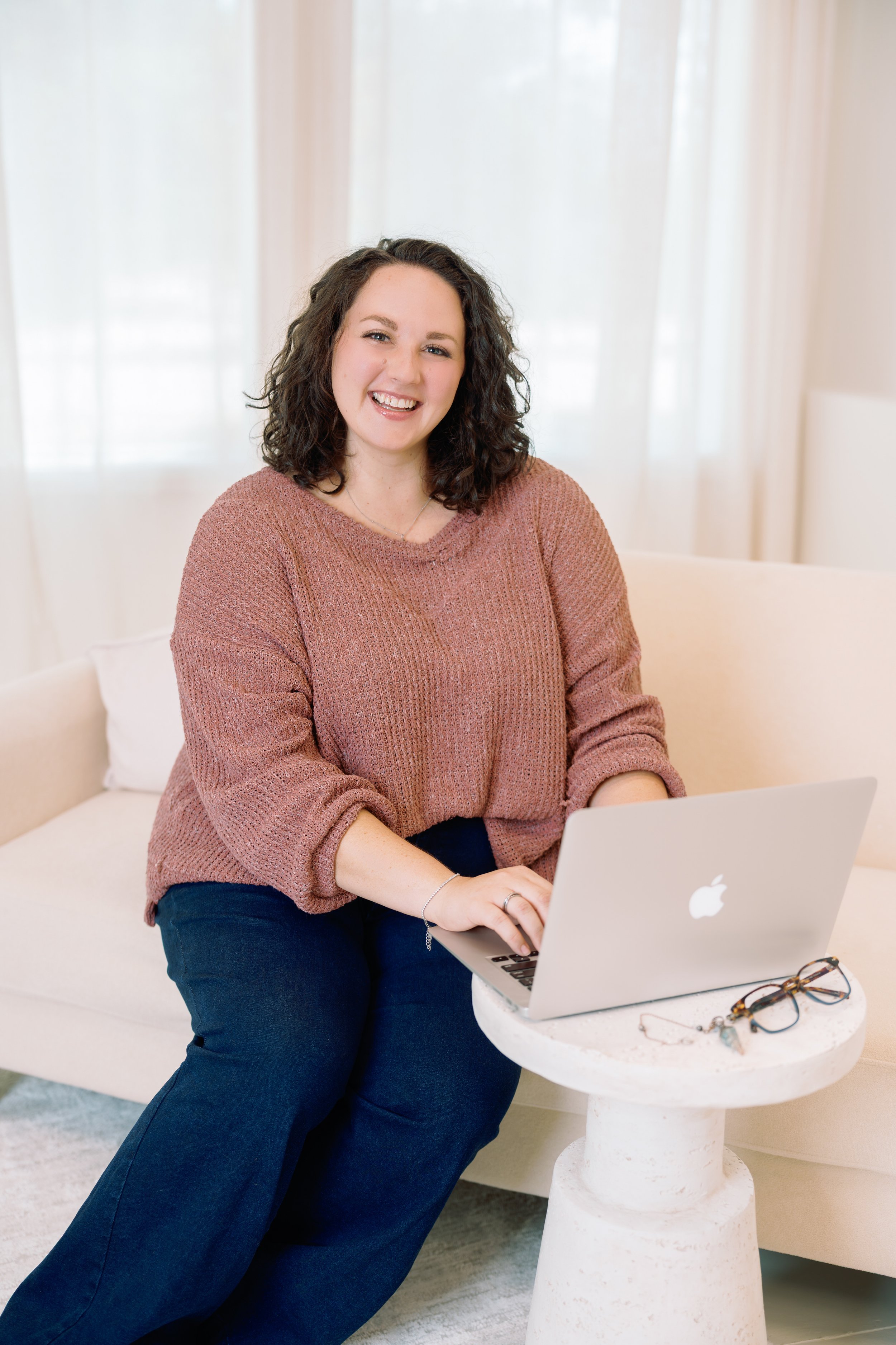 A woman with curly brown hair smiling while using a silver MacBook at a small white round table. She is wearing a pinkish-brown sweater and dark blue jeans, with glasses and jewelry nearby. The background features soft, sheer curtains and a light-colored interior.