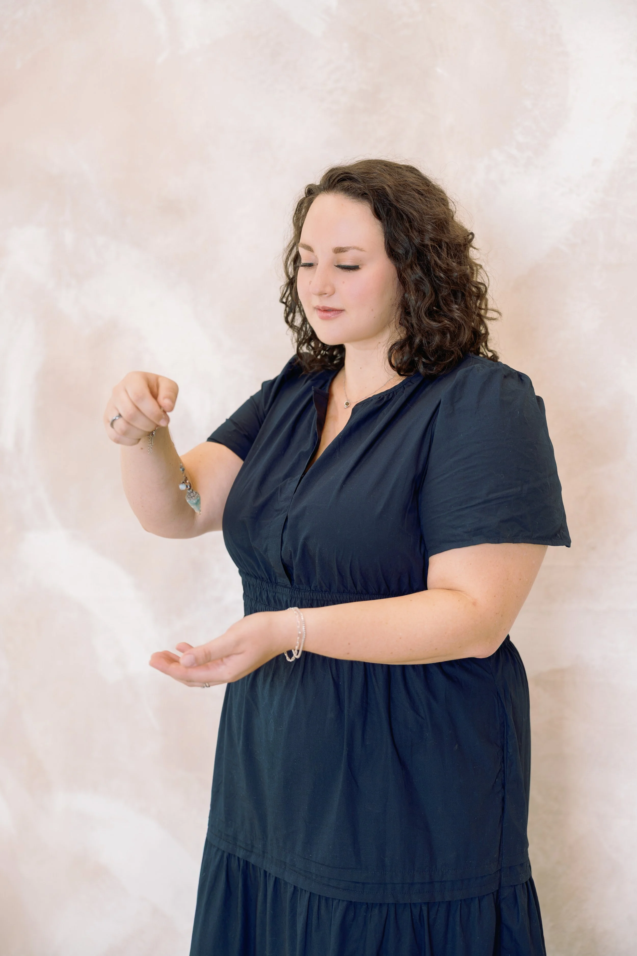 A woman with curly brown hair wearing a dark blue dress, holding a necklace with a large crystal pendant, standing against a light-colored wall.