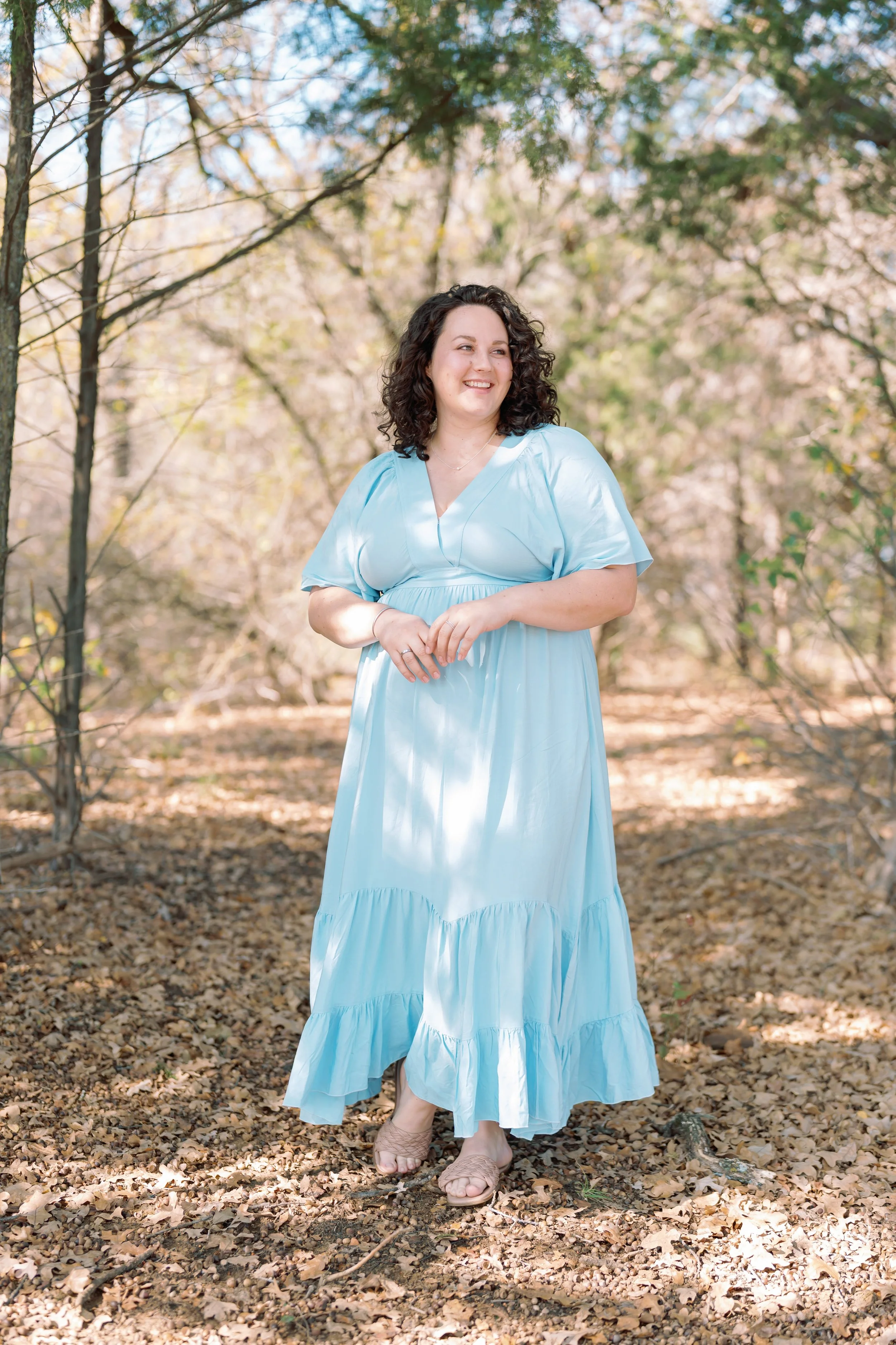 A woman with curly dark hair wearing a light blue dress and sandals standing in a wooded area with fallen leaves.