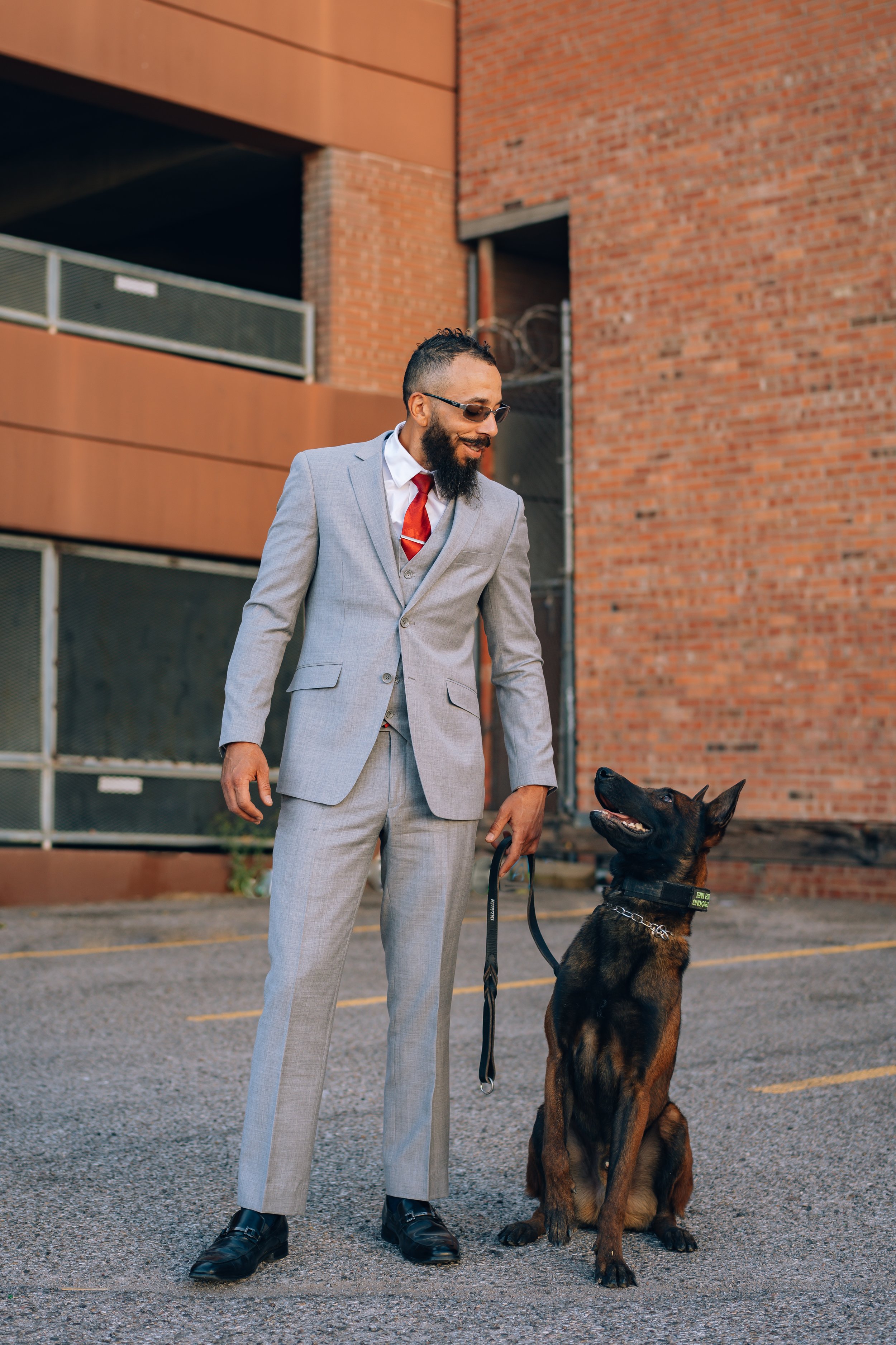 A man in a light gray suit with a red tie and sunglasses stands outside holding a leash attached to a large German Shepherd dog. They are in a parking lot with a brick building in the background.