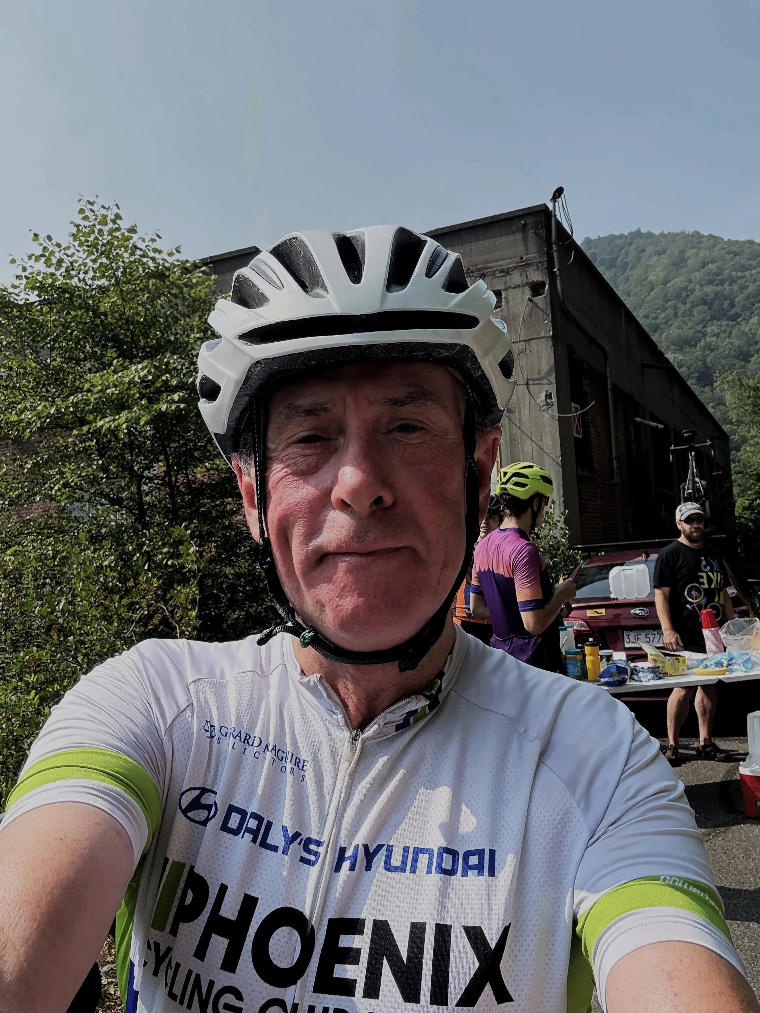 A man in a white cycling jersey and helmet taking a selfie outdoors, with people, a table, and a car in the background.