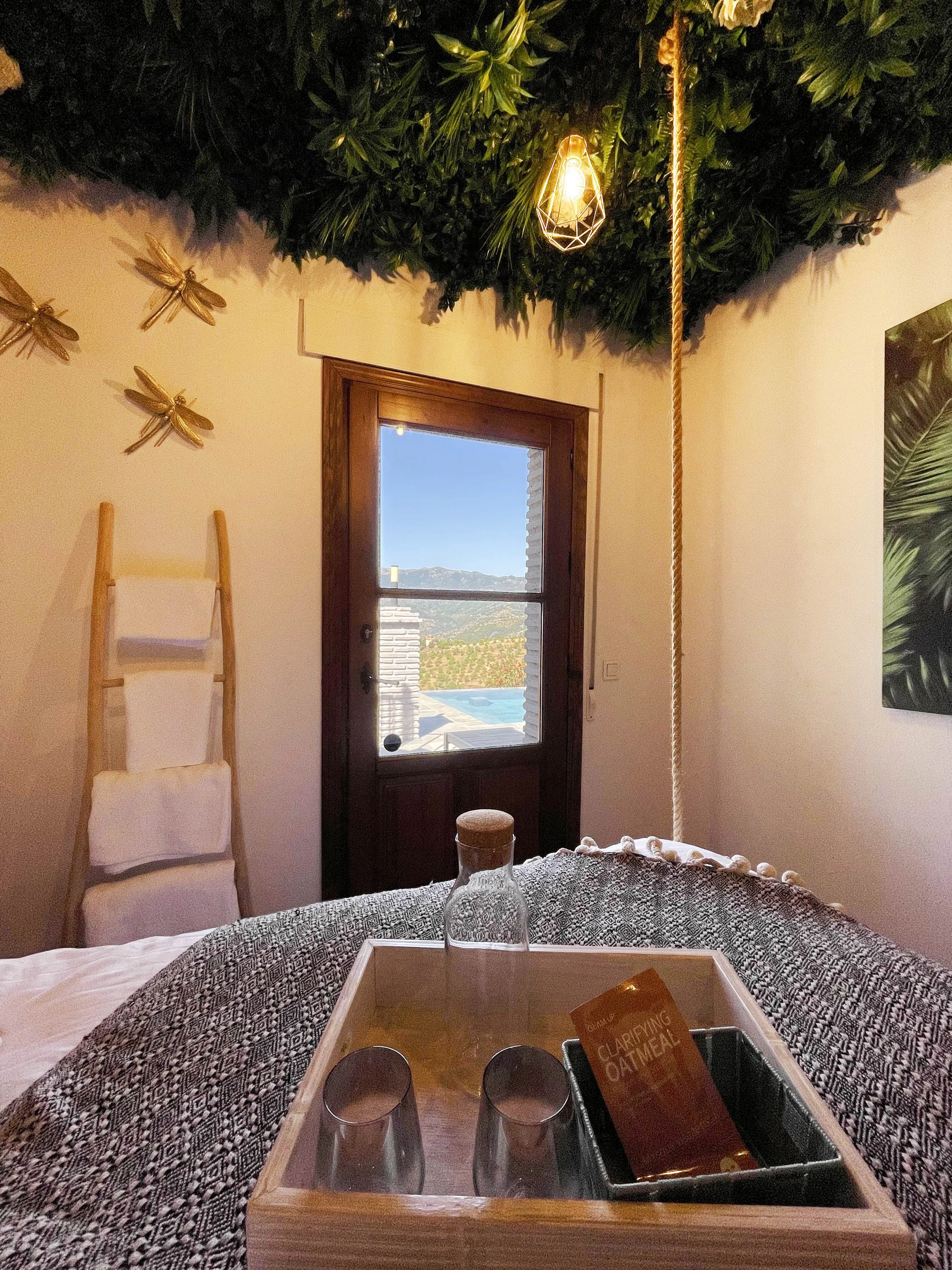Interieur van een slaapkamer met een raam dat uitzicht biedt op een zwembad en heuvels in de verte. Boven het bed hangt een groene vegetatie aan het plafond met een hangende lamp. Aan de muur hangen decoratieve gouden vliegen en een houten ladder. Op het bed ligt een bord met de tekst "Clarifying Oatmeal" en twee glazen.