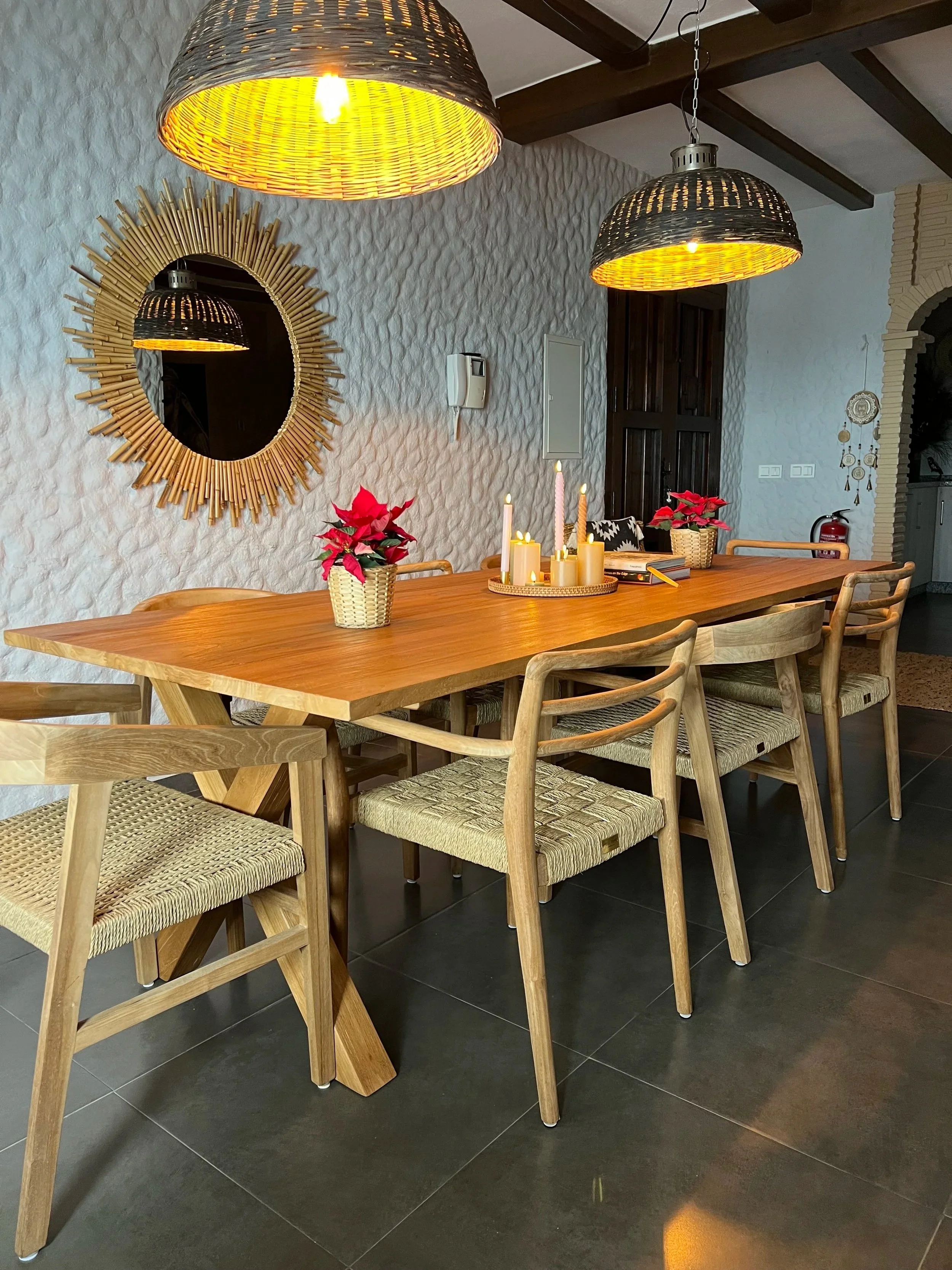 Een eetkamer met een houten tafel en tuinmeubilair, plafond met houten balken, hanglampen en een keuken in de achtergrond.