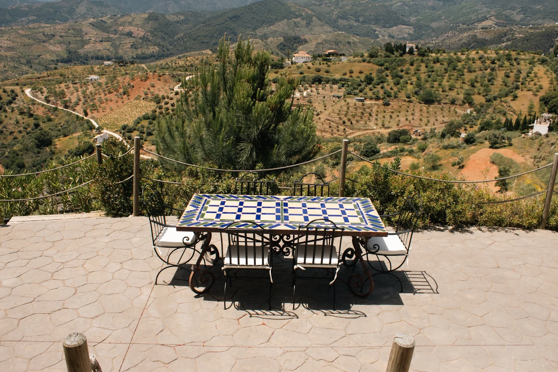 Tafel met kleurrijk patroon op een terras met uitzicht op heuvelachtig landschap met verspreide huizen en groene olijf- en dennenbomen.