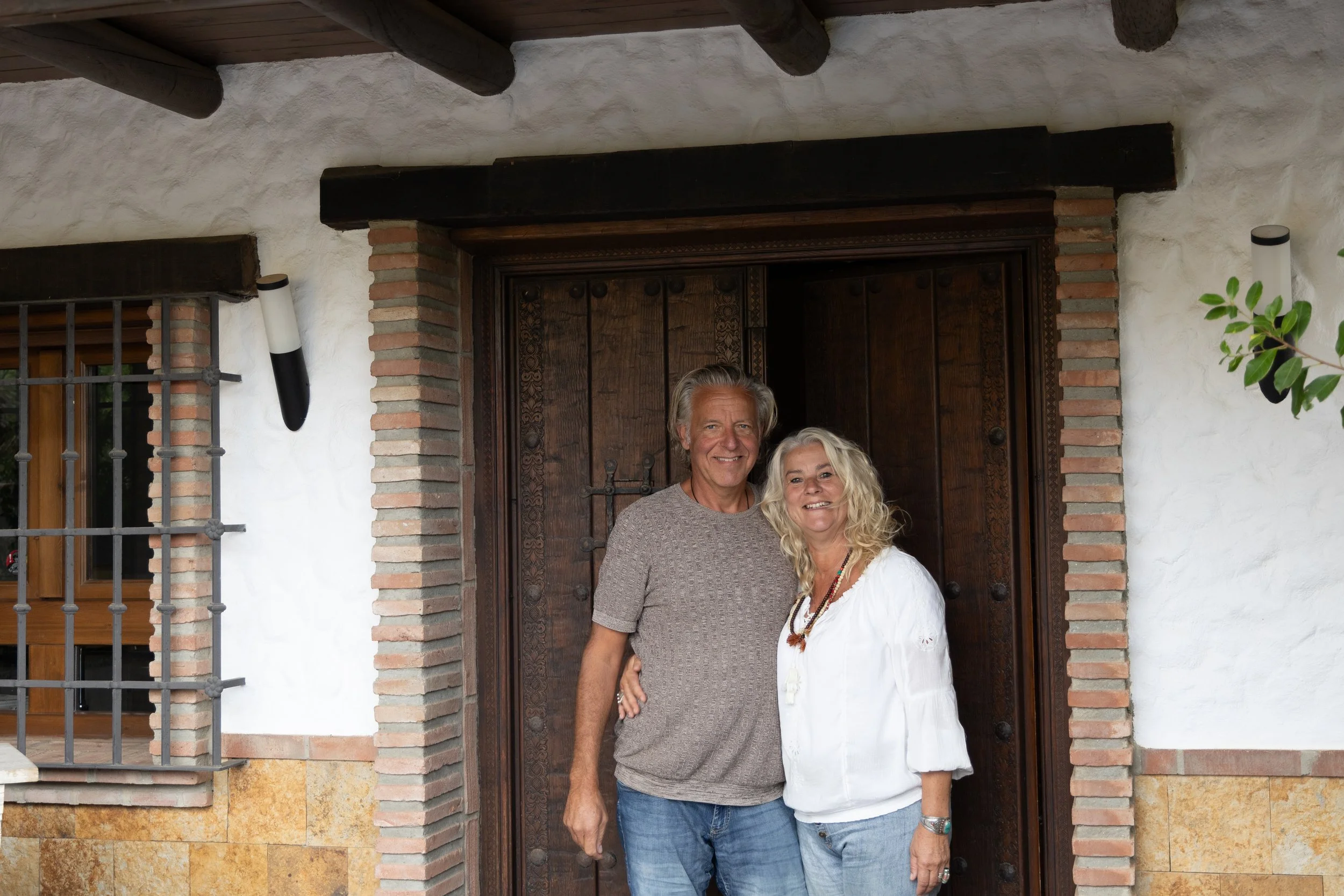 Een oudere man en vrouw staan samen voor een houten deur in een witte muur met stenen en bakstenen accenten.