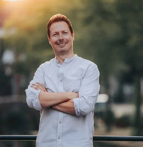 Man met gekruiste armen, lachend, buiten met groene bomen op de achtergrond.