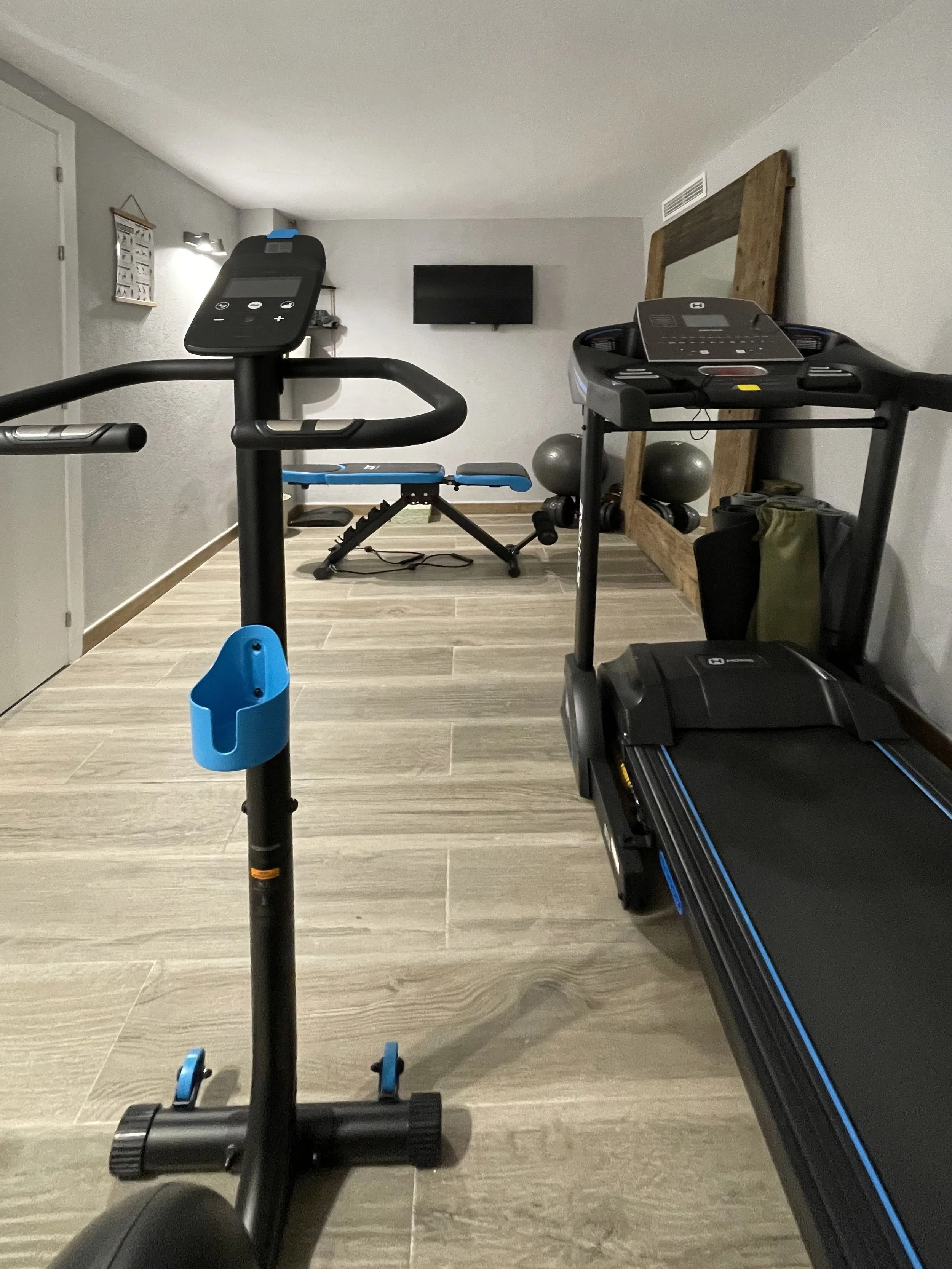 Een binnenruimte met fitnessapparatuur, waaronder een hometrainer, een loopband en een gewichtentafel, met een muur en een televisie aan de achtergrond.