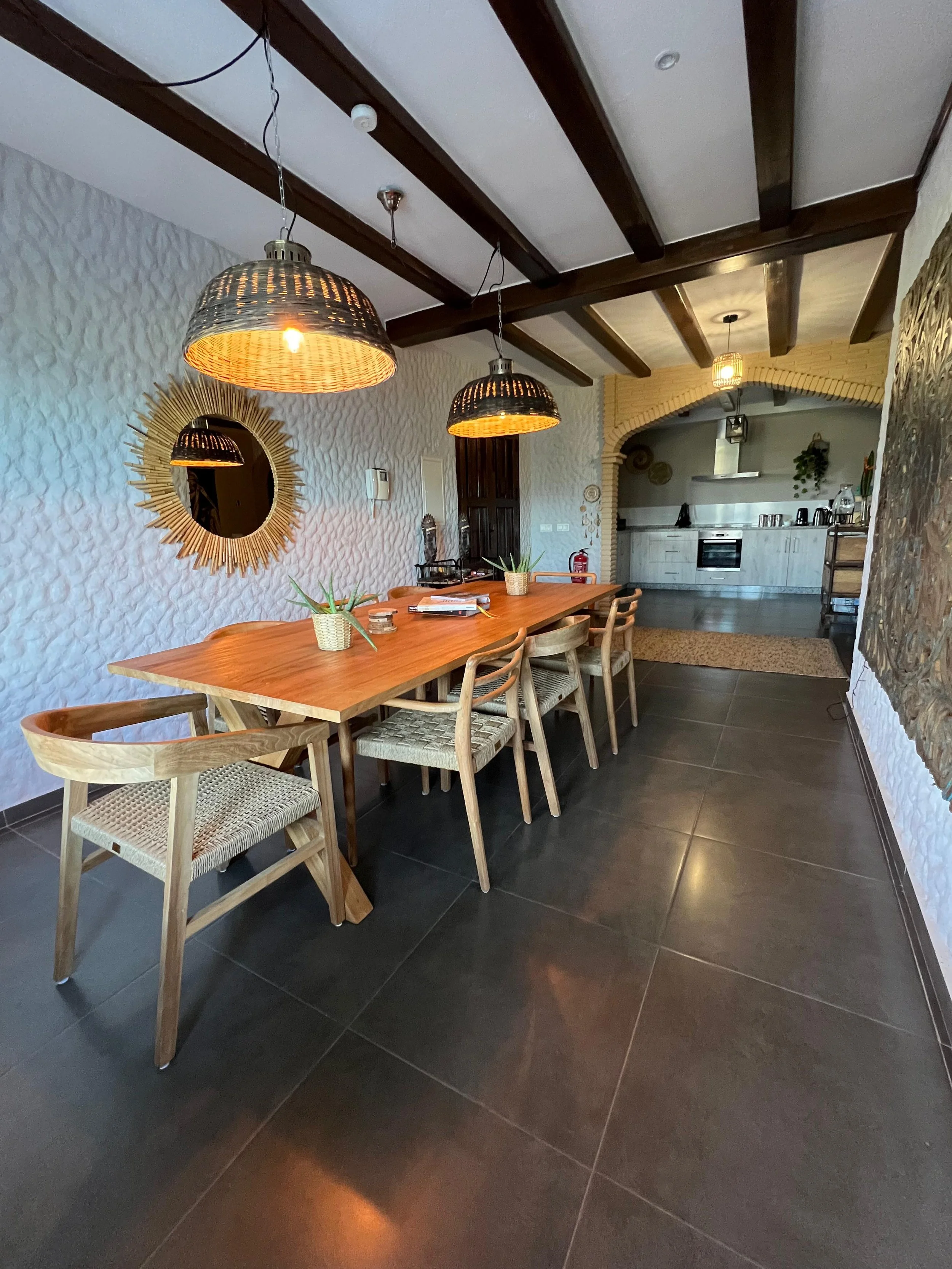Binnenruimtes met een houten tafel, houten stoelen, drie hanglampen, een ronde spiegel met een zonnestraalrand, een keuken in de achtergrond en decoraties op de muur.
