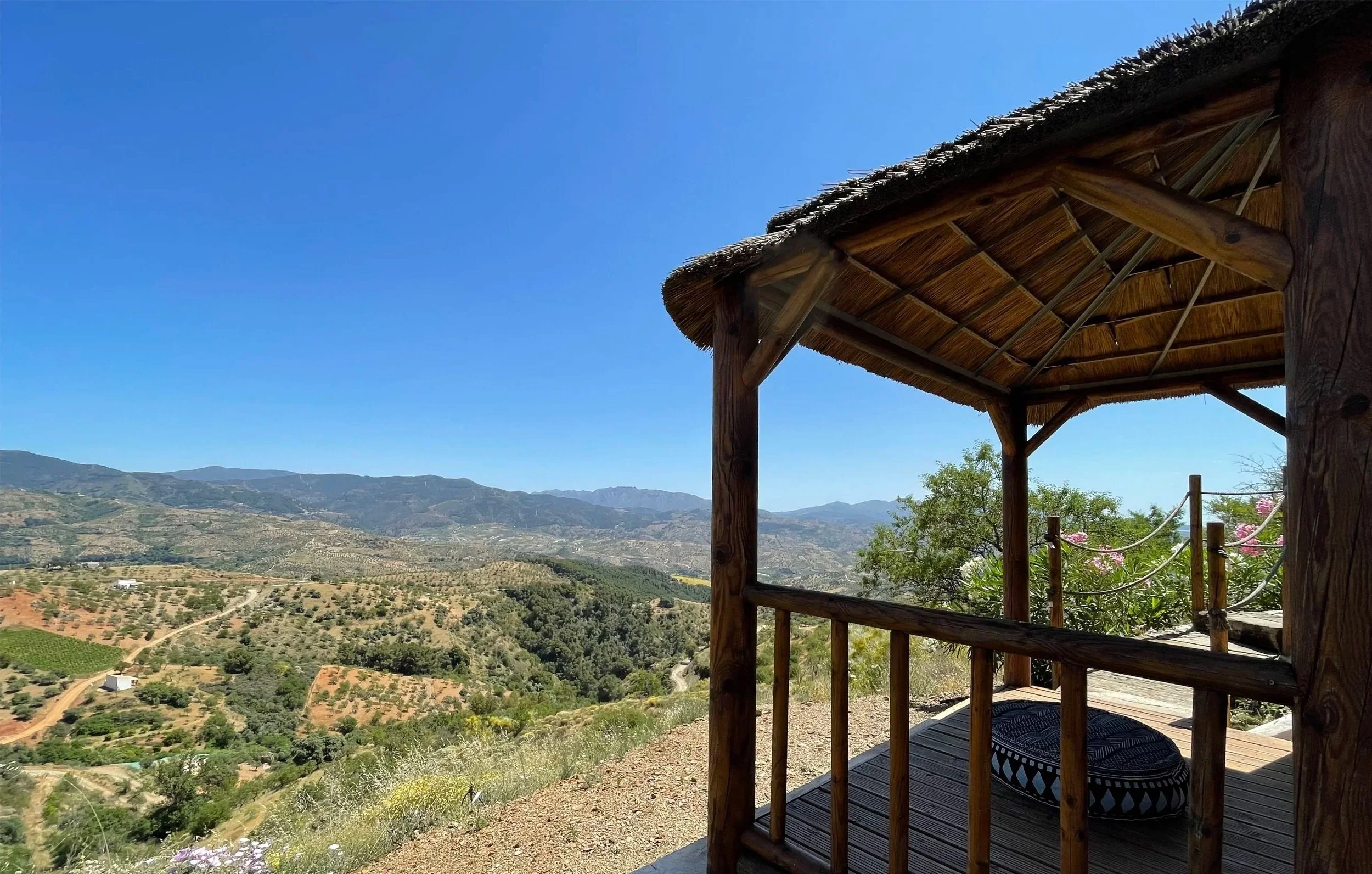 Bergachtig landschap met een houten terras of uitzichtplatform onder een dat van stro gemaakte overkapping, uitzicht op bergen en valleien onder een heldere blauwe hemel.